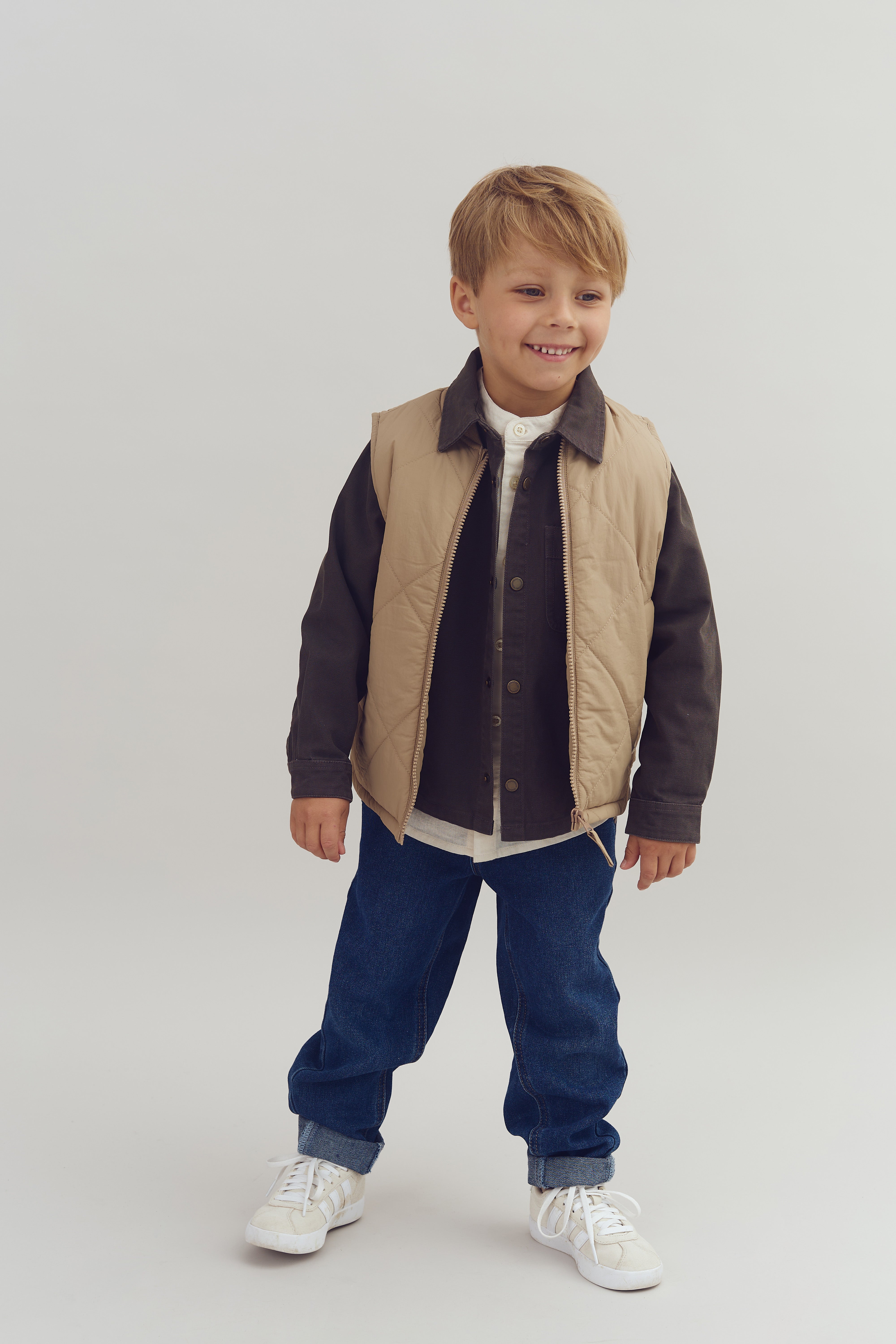 Dreng smiler, iført beige vest, mørk skjorte og blå jeans. Han står på en simpel hvid baggrund.
