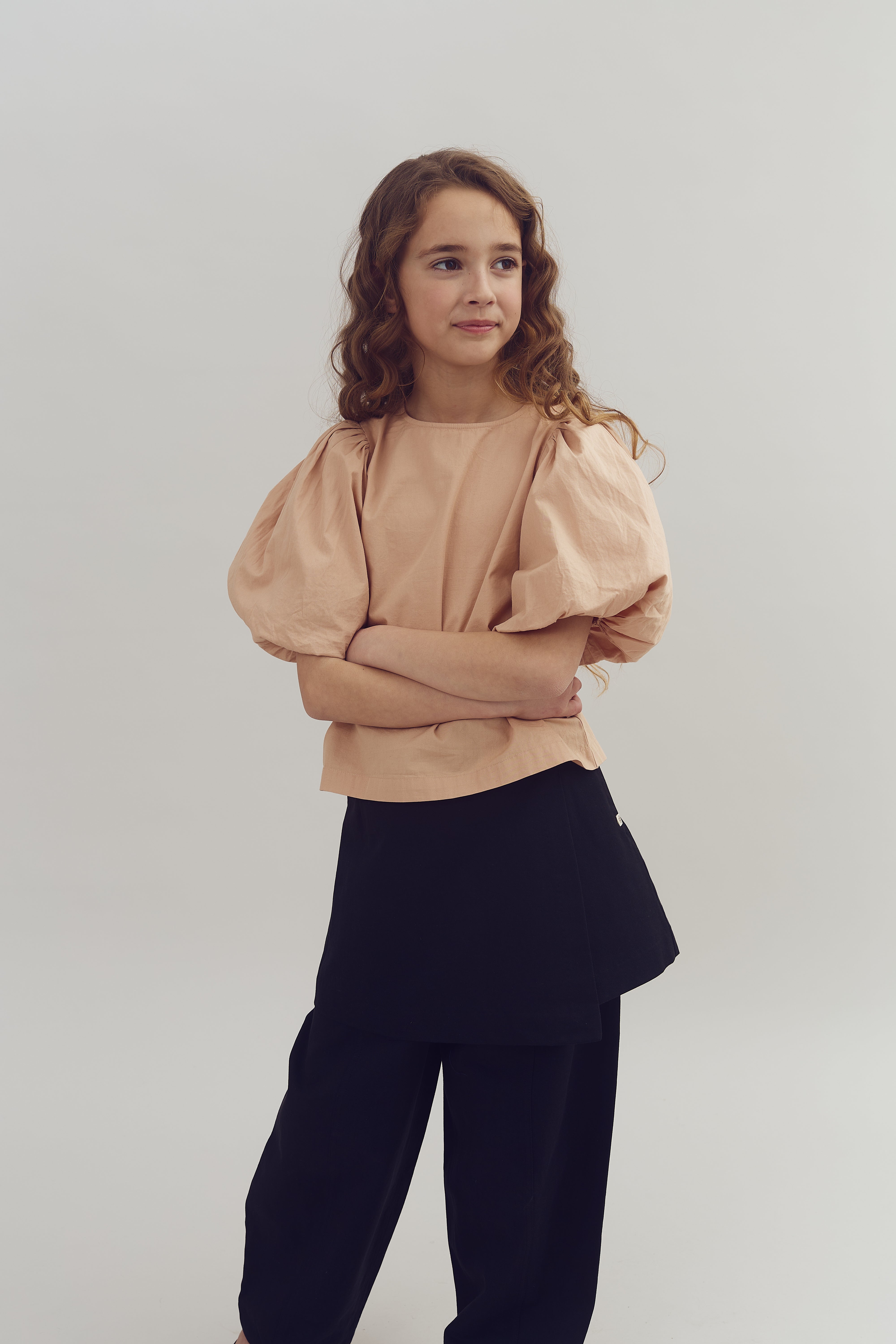 En ung pige står med korslagte arme, iført en beige bluse med puffede ærmer og sorte bukser, i et neutralt studio miljø.