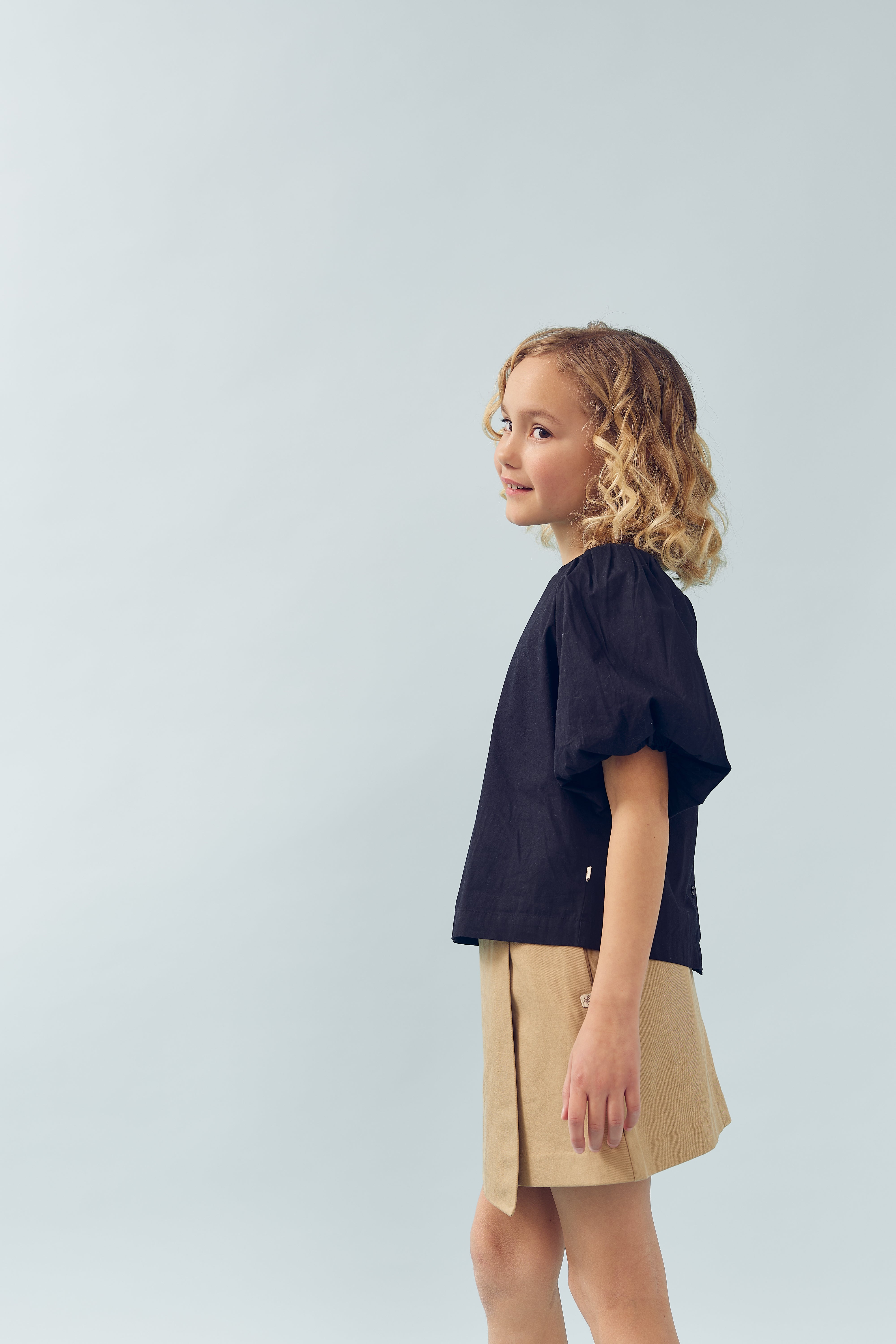 Et barn med krøllet hår står i profil. Barnet bærer en sort bluse og beige nederdel, mod en lys baggrund i et studieagtigt miljø.