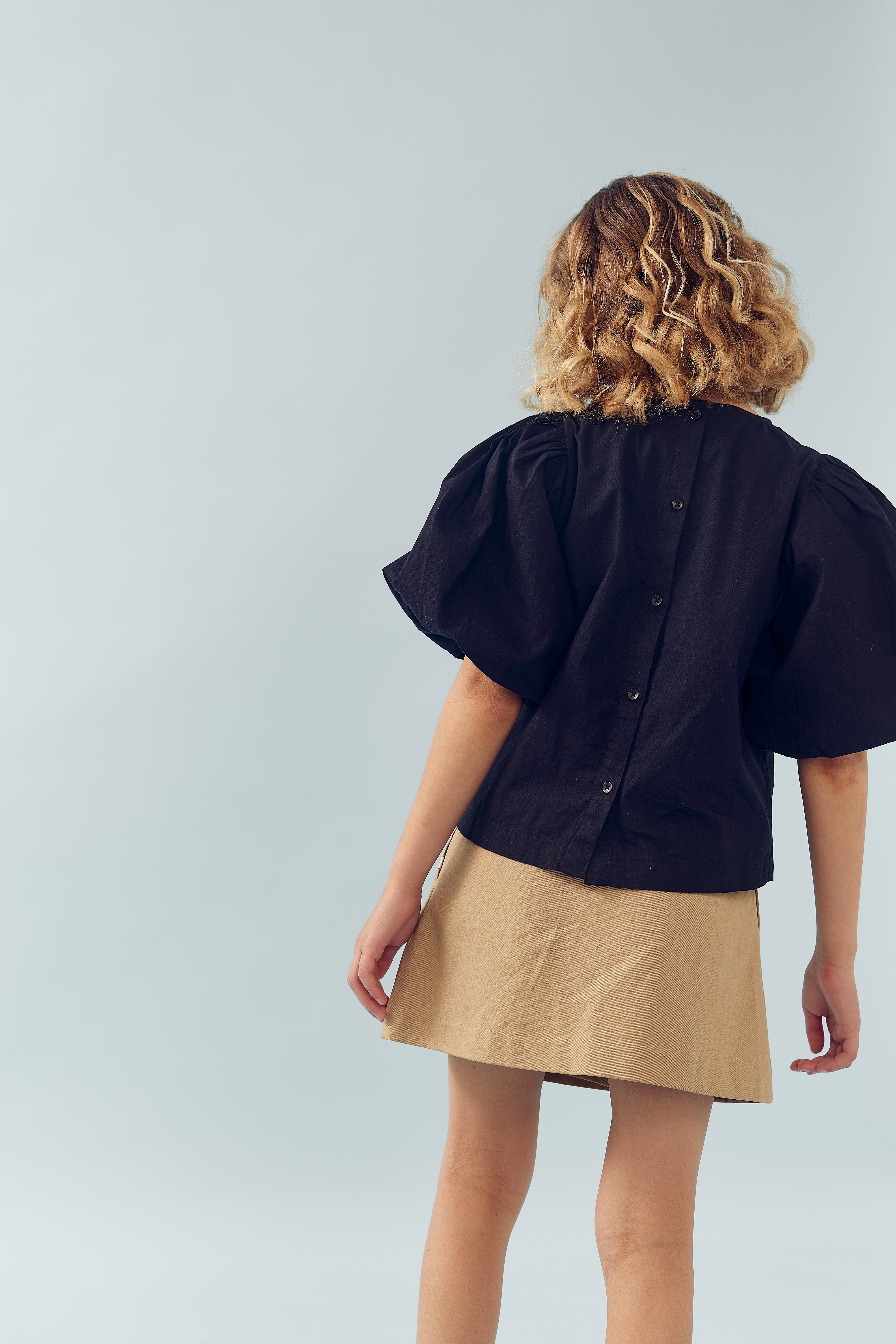 Person står med ryggen til iført en sort bluse med knapper og en beige nederdel. Kontexten er en simpel, lys baggrund.