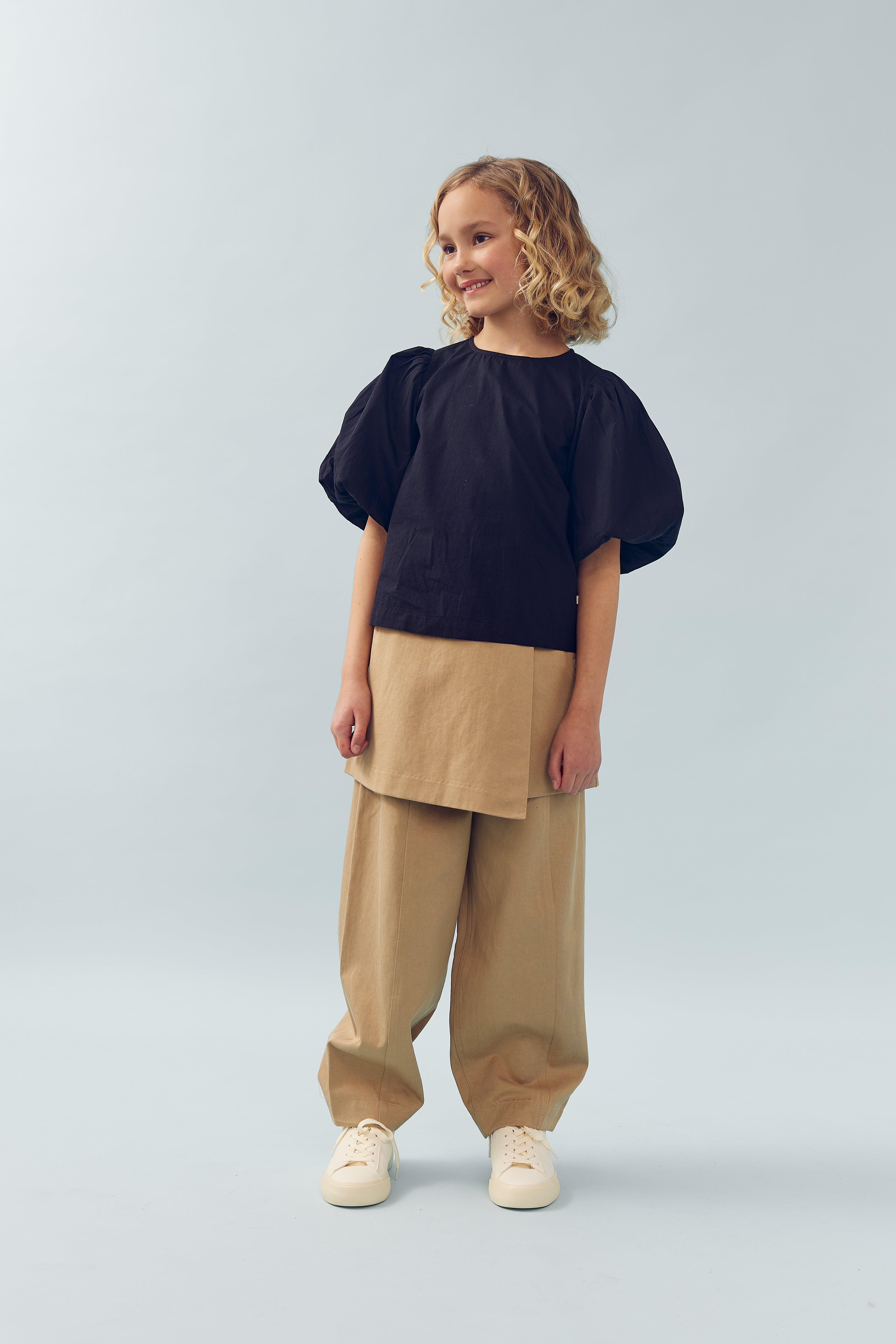 Barn står smilende iført en sort bluse med pufærmer og beige bukser. Baggrundens lyseblå farve skaber en rolig atmosfære.