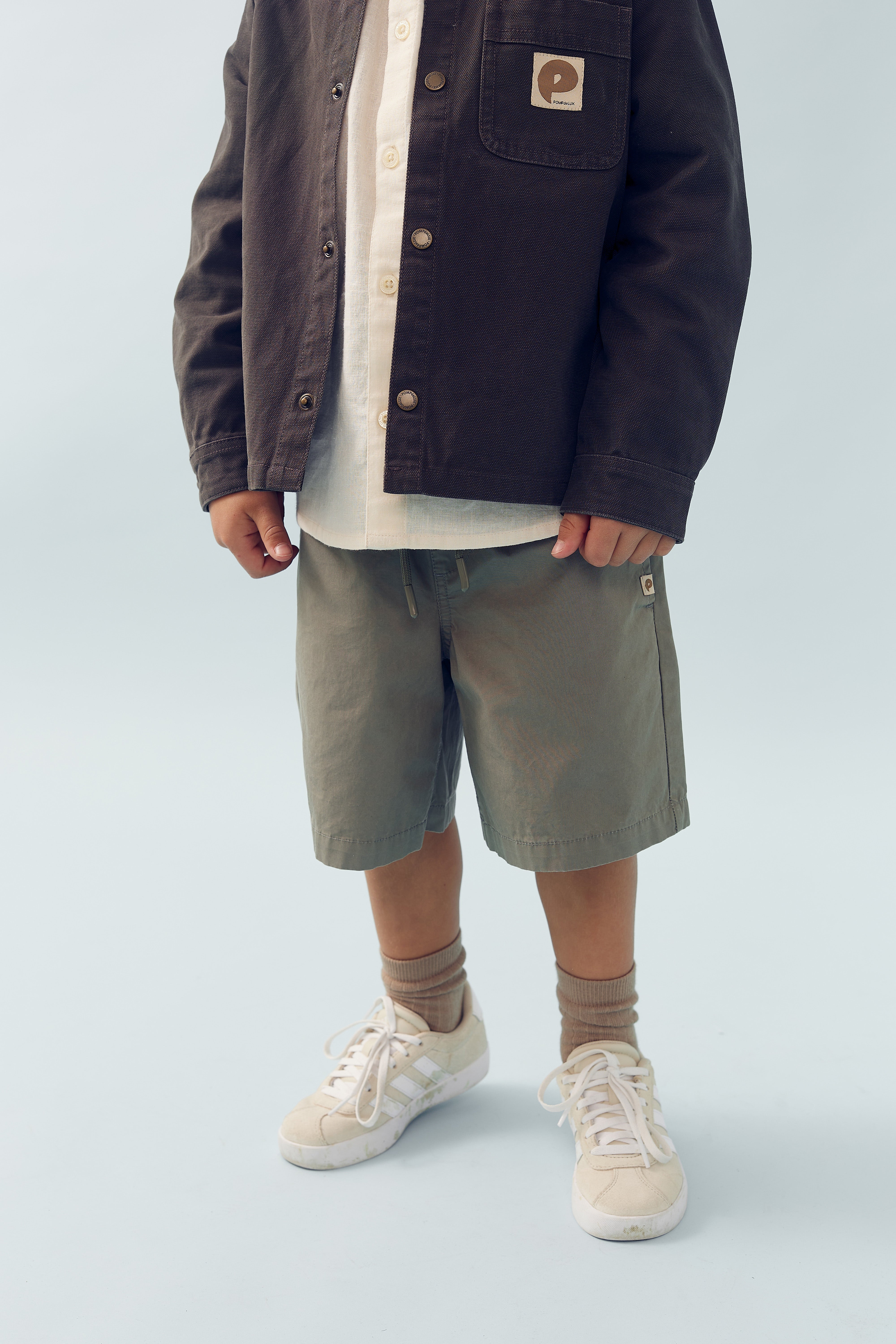 Et barn bærer en jakke og shorts og står stille. De har sneakers og sokker på. Lommen på jakken har et mærke med et logo i et minimalistisk studieområde.