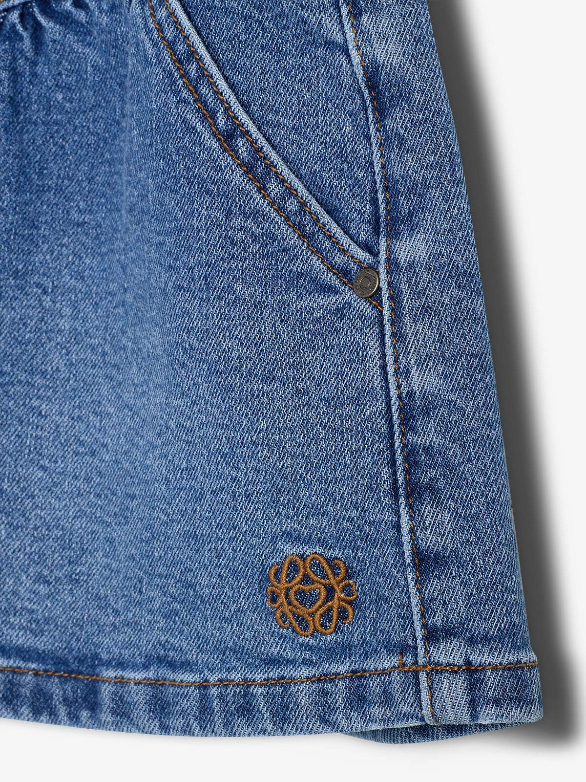 A denim fabric with a pocket, featuring orange stitching and a small embroidered symbol near the bottom edge, possibly a decorative pattern, on a plain background.
