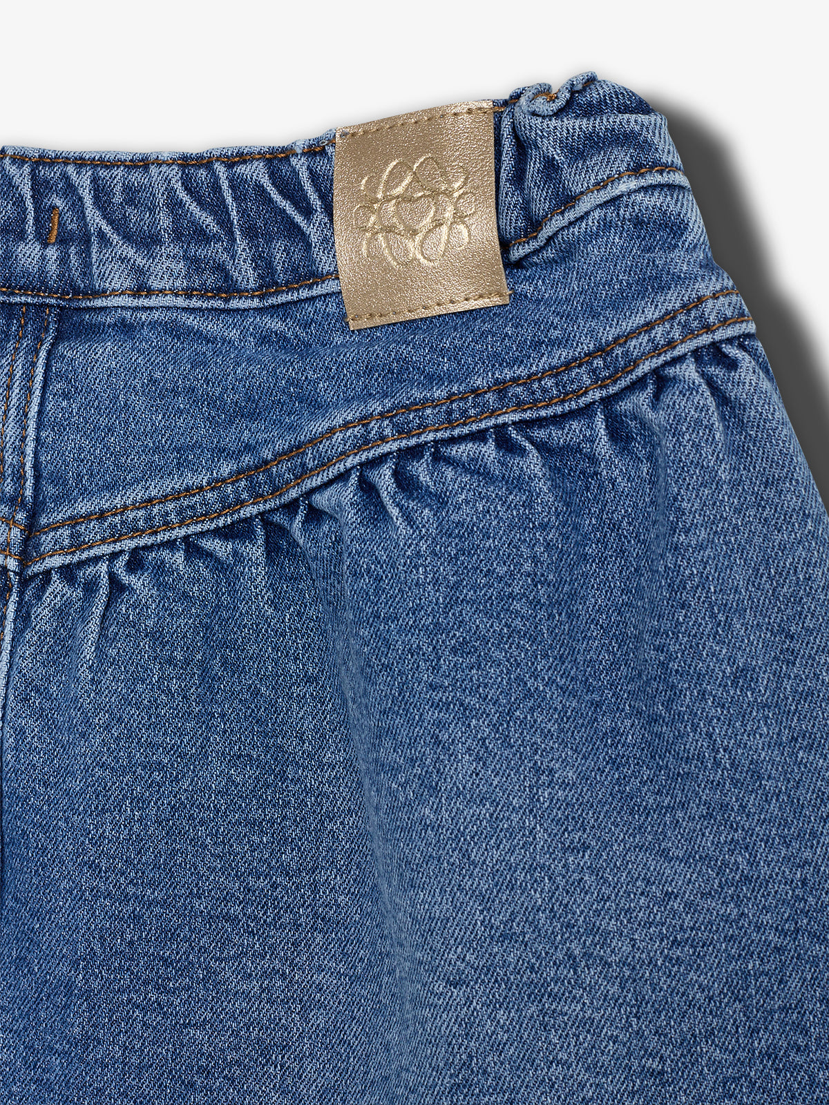 Jeansmateriale med en elastisk linning, hvor der er placeret en beige læderlap med et præget blomstermønster øverst. Baggrunden er lys og ensfarvet.