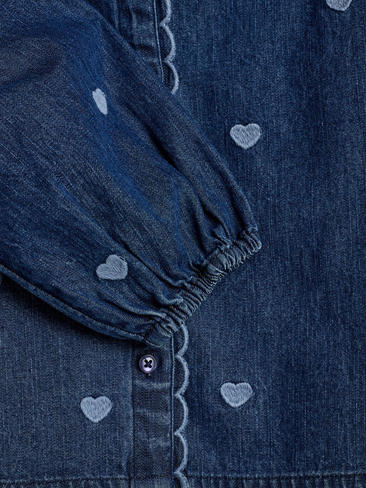 En mørkeblå denimskjorte med lysere hjertebroderi, knappelukning og elastiske ærmer. Stoffet er detaljeret med bølgede kanter langs knaplukningen og hele ærmet.