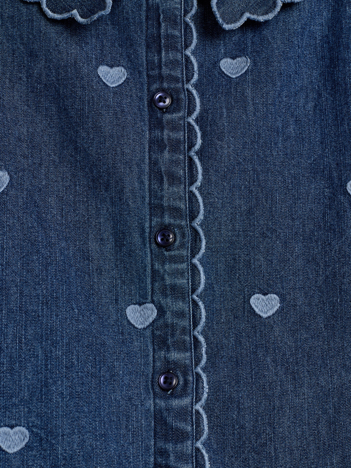 Blå denimskjorte med hjertebroderi. Skjorten har en bølget kant langs knapstolpen med mørke knapper. Hjertemønsteret tilføjer et blødt, dekorativt element.