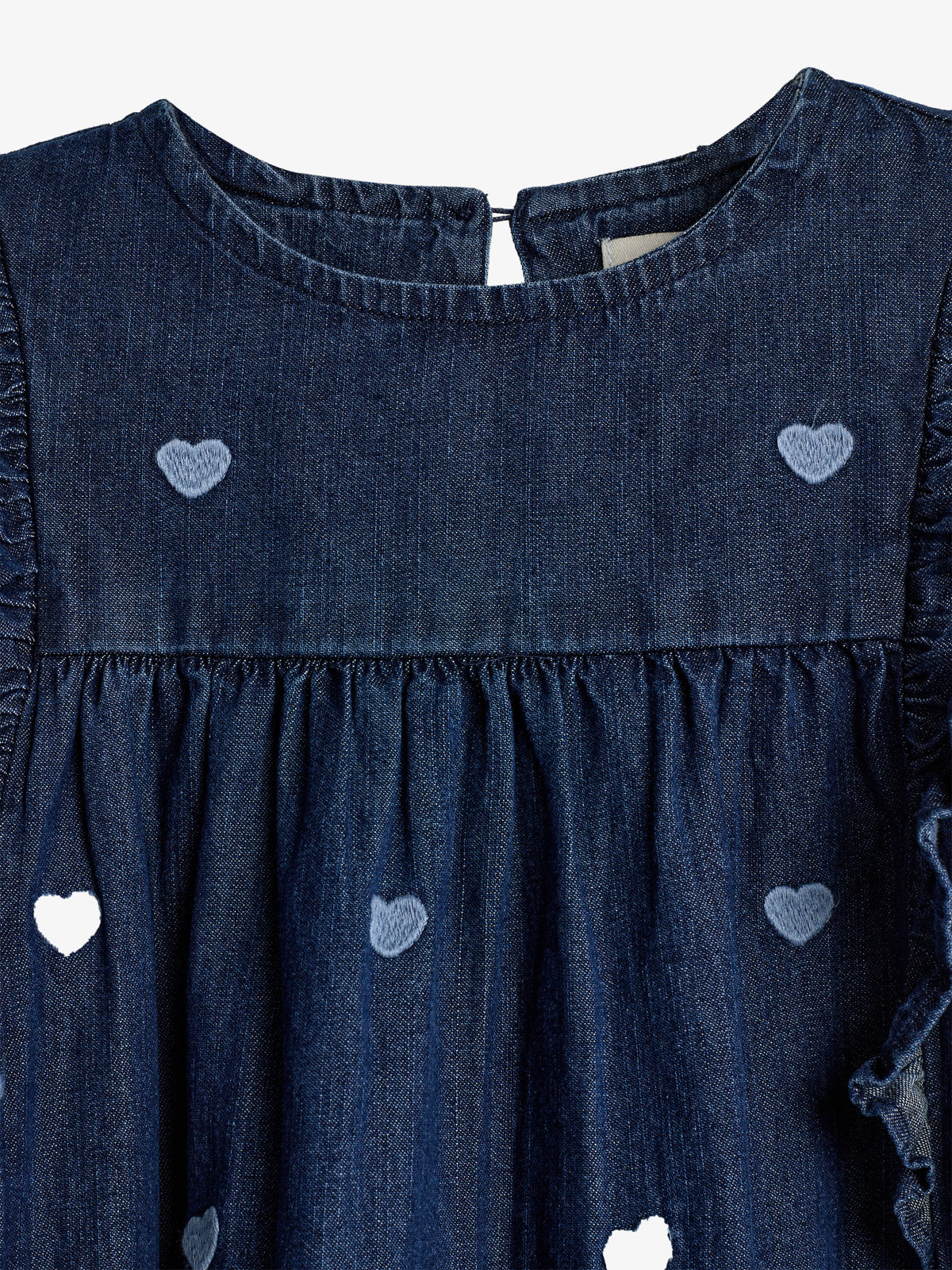 Kjole af denim med hjertebroderier i lyseblå farve. Kjolen har en rund halsudskæring og små flæser ved ærmerne. Baggrunden er ensfarvet lysegrå. Ingen tekst er synlig.
