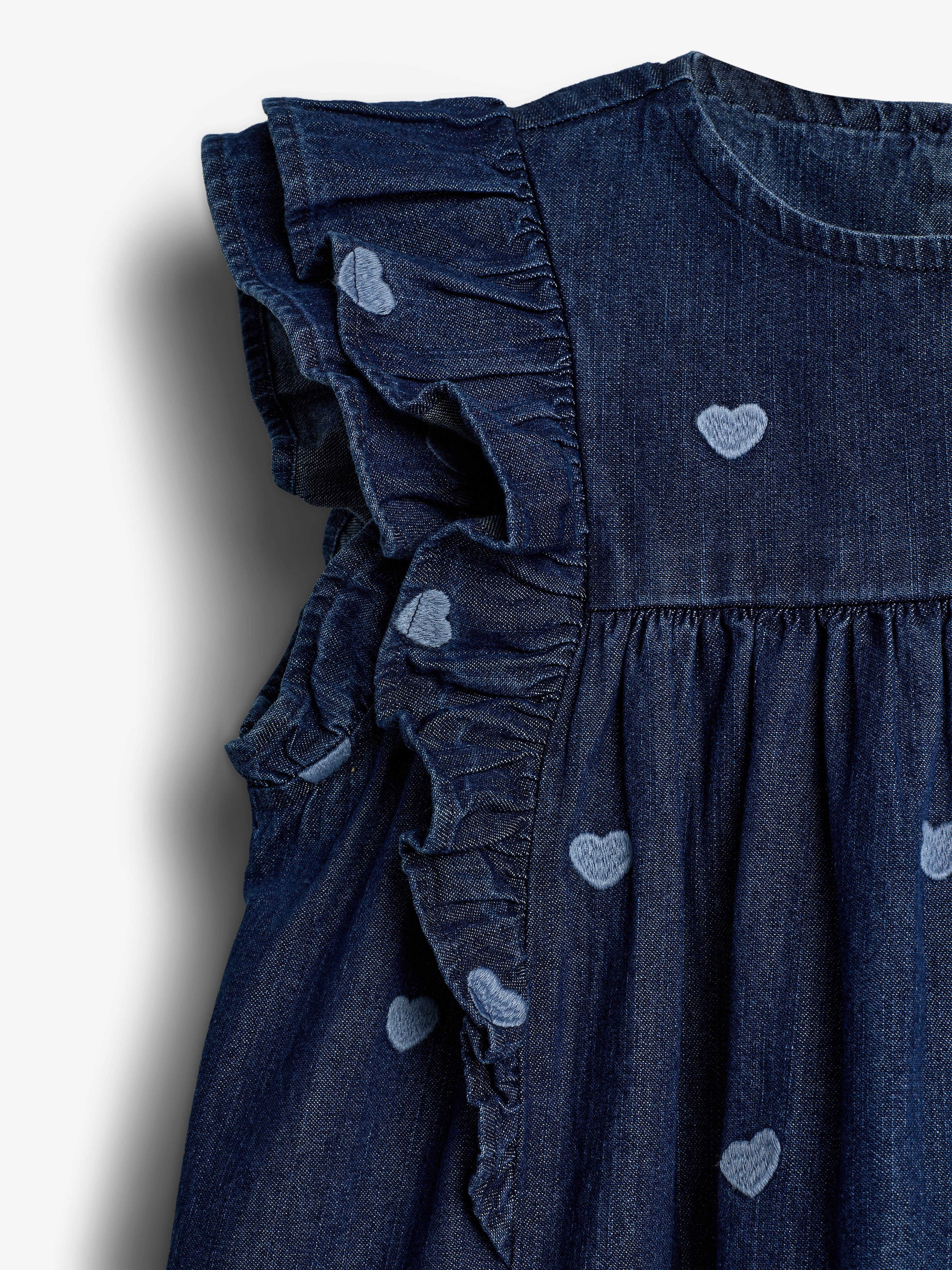 Blå denimkjole med flæser og lyseblå hjerteprint, der pynter stoffet. Kjolen hænger mod en lys baggrund, der fremhæver materialets tekstur og detaljer.