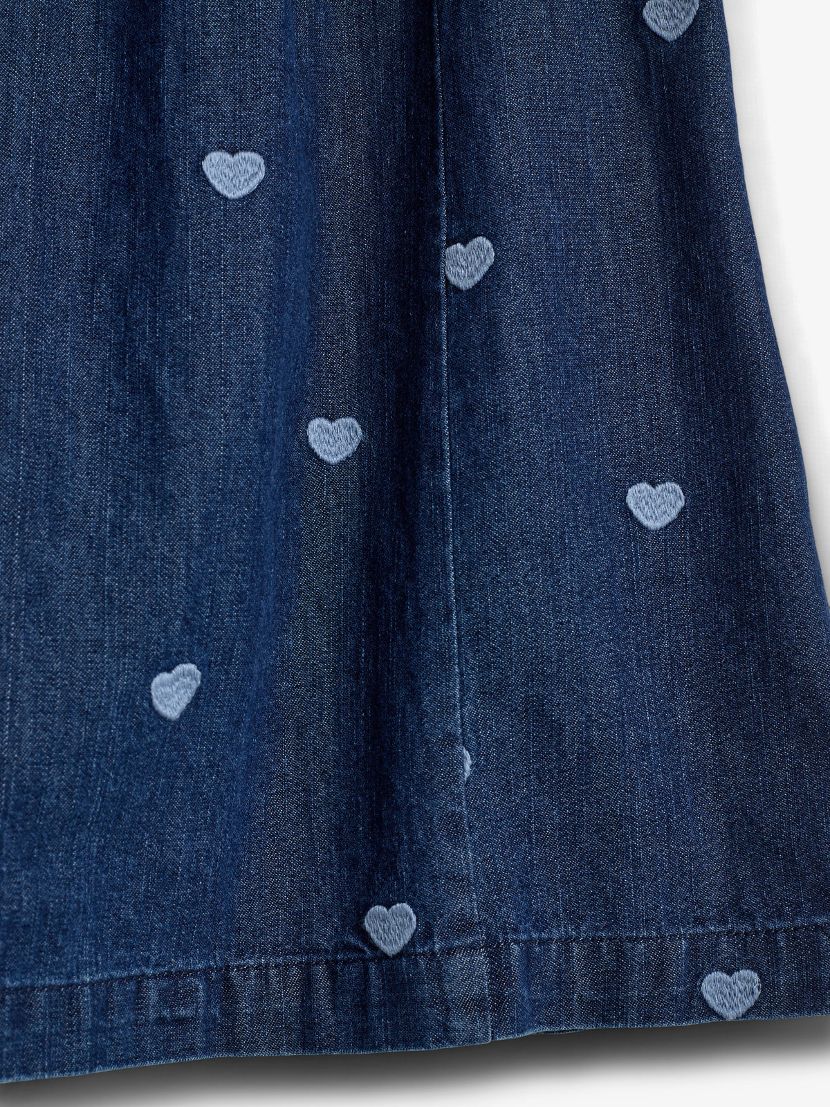 Blå denimstof med små, lyseblå broderede hjerter fordelt jævnt på overfladen. Stoffet hænger vertikalt i et neutralt lys uden yderligere dekorationer.