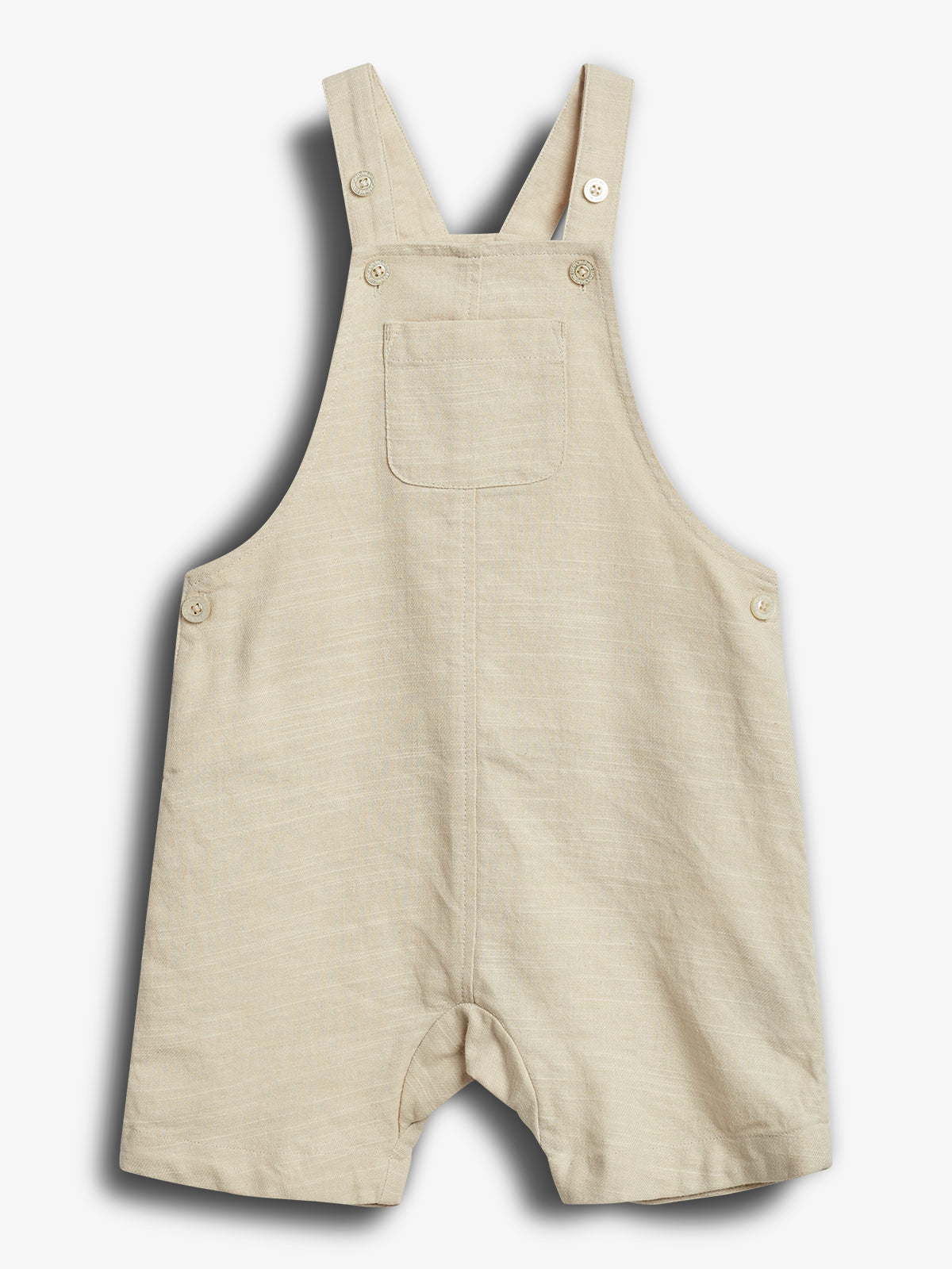 Beige kort overalls med lommer foran, knapper på stropperne og siderne, hænger fladt mod en lys baggrund.