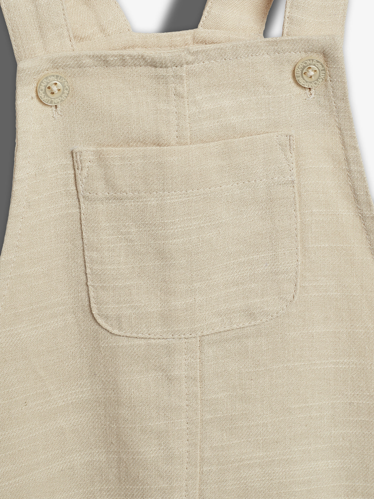 Et nærbillede af en lys beige overall viser en brystlomme med to knapper over den. Knapperne er præget med teksten "BONPOINT". Baggrunden er enkel og grå.