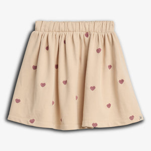 En beige nederdel med elastisk talje og små pink hjerteprint spredt over stoffet. Baggrunden er ensfarvet lys grå, hvilket fremhæver nederdelens detaljerede design.