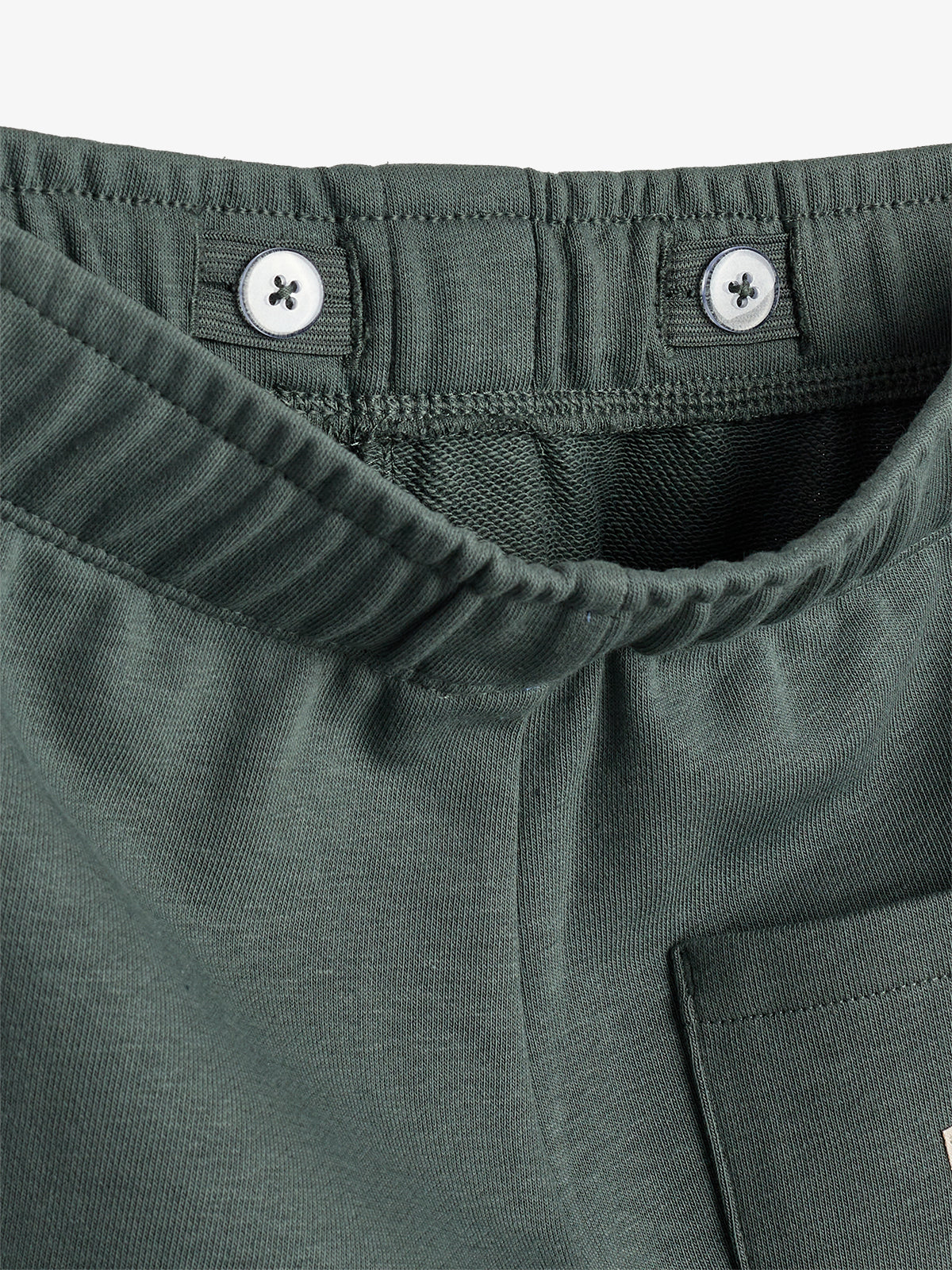 Green elastic waistband with buttons is on a pair of pants, featuring ribbed fabric and a patch pocket. The focus is on the waistband's stitching and button details.