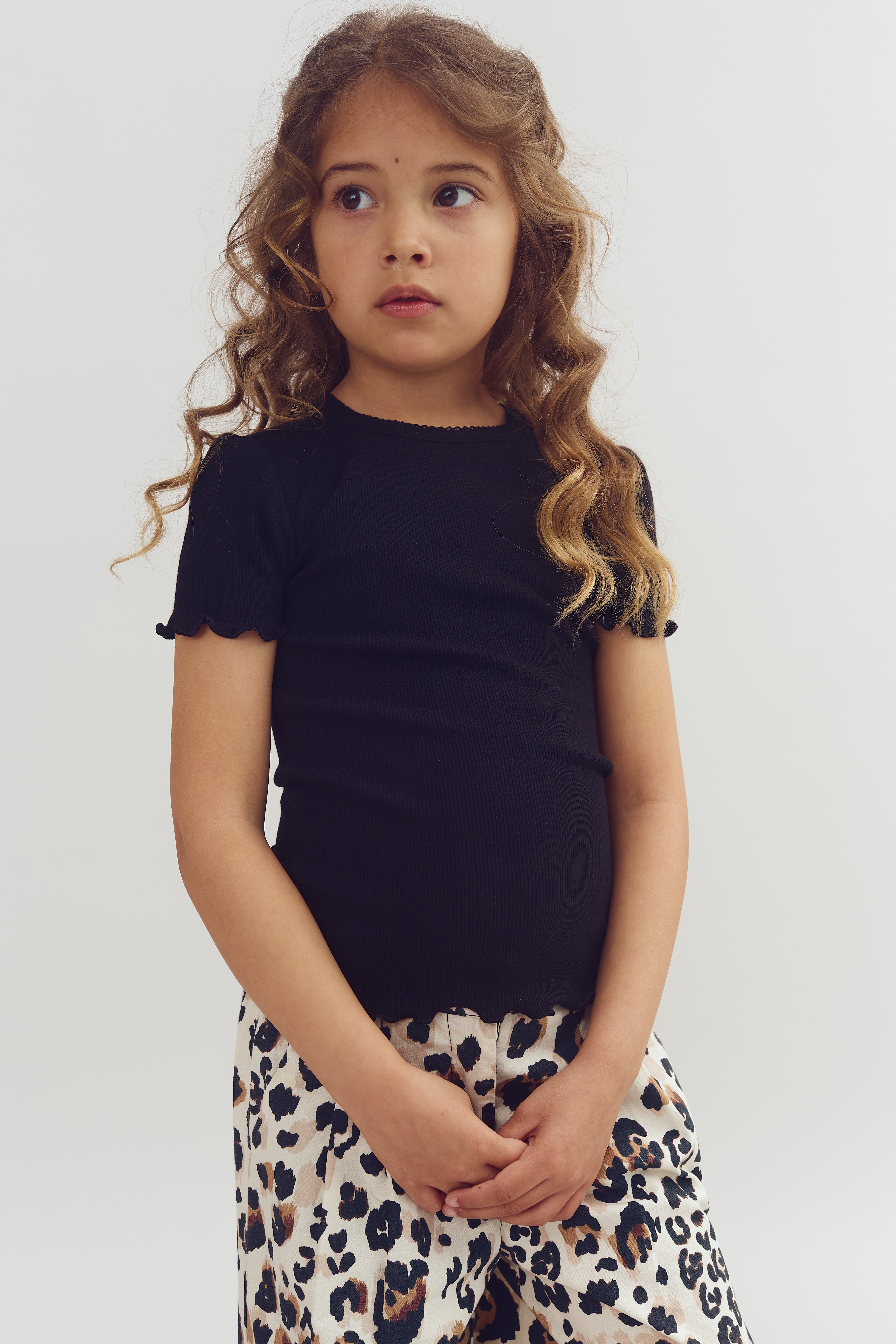 A young girl with long curly hair stands still, wearing a black shirt and leopard-print pants, against a plain light-colored background. She's looking slightly to the side.