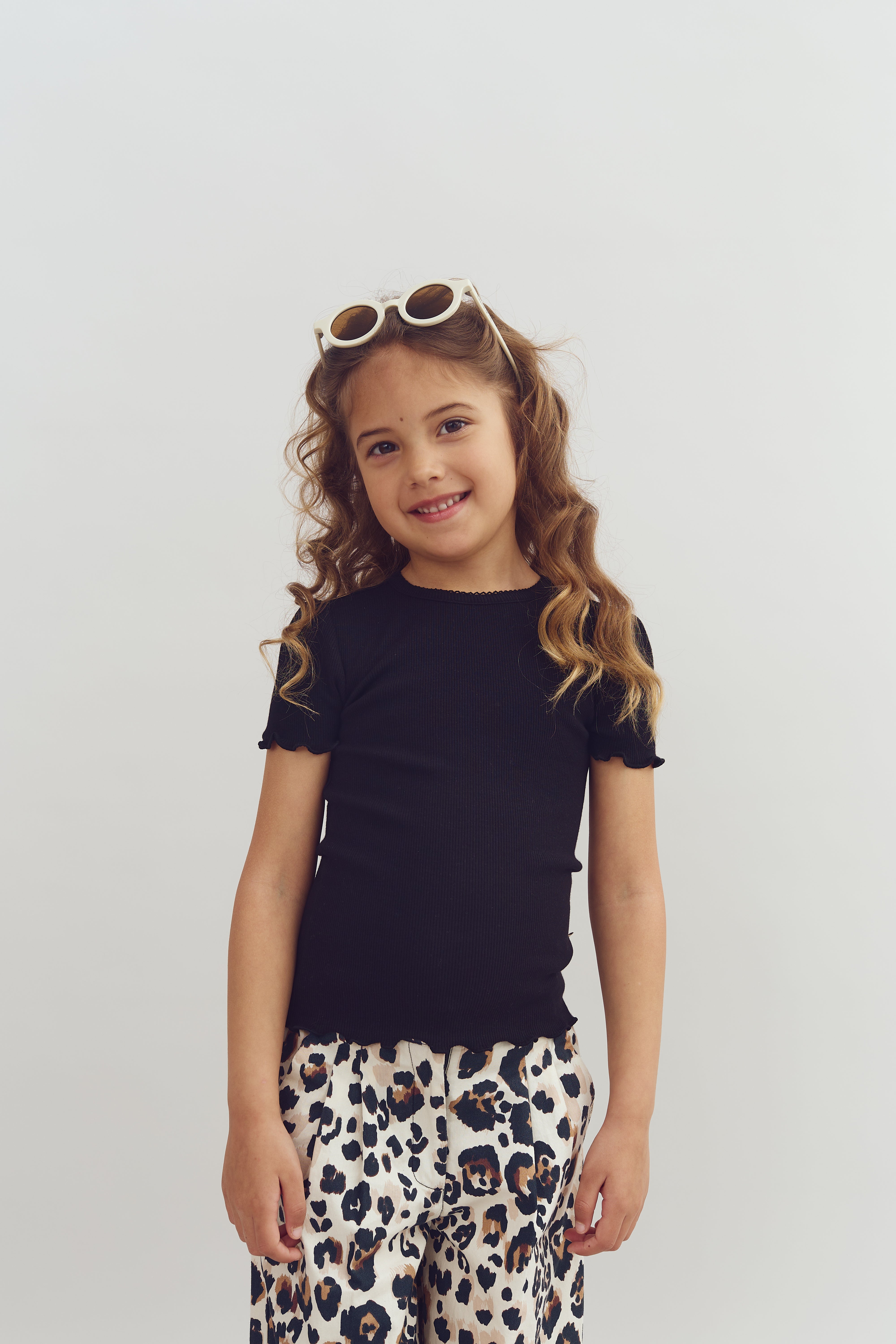 A child stands smiling, wearing a black shirt and leopard print pants, with sunglasses resting on their head, against a plain, light background.