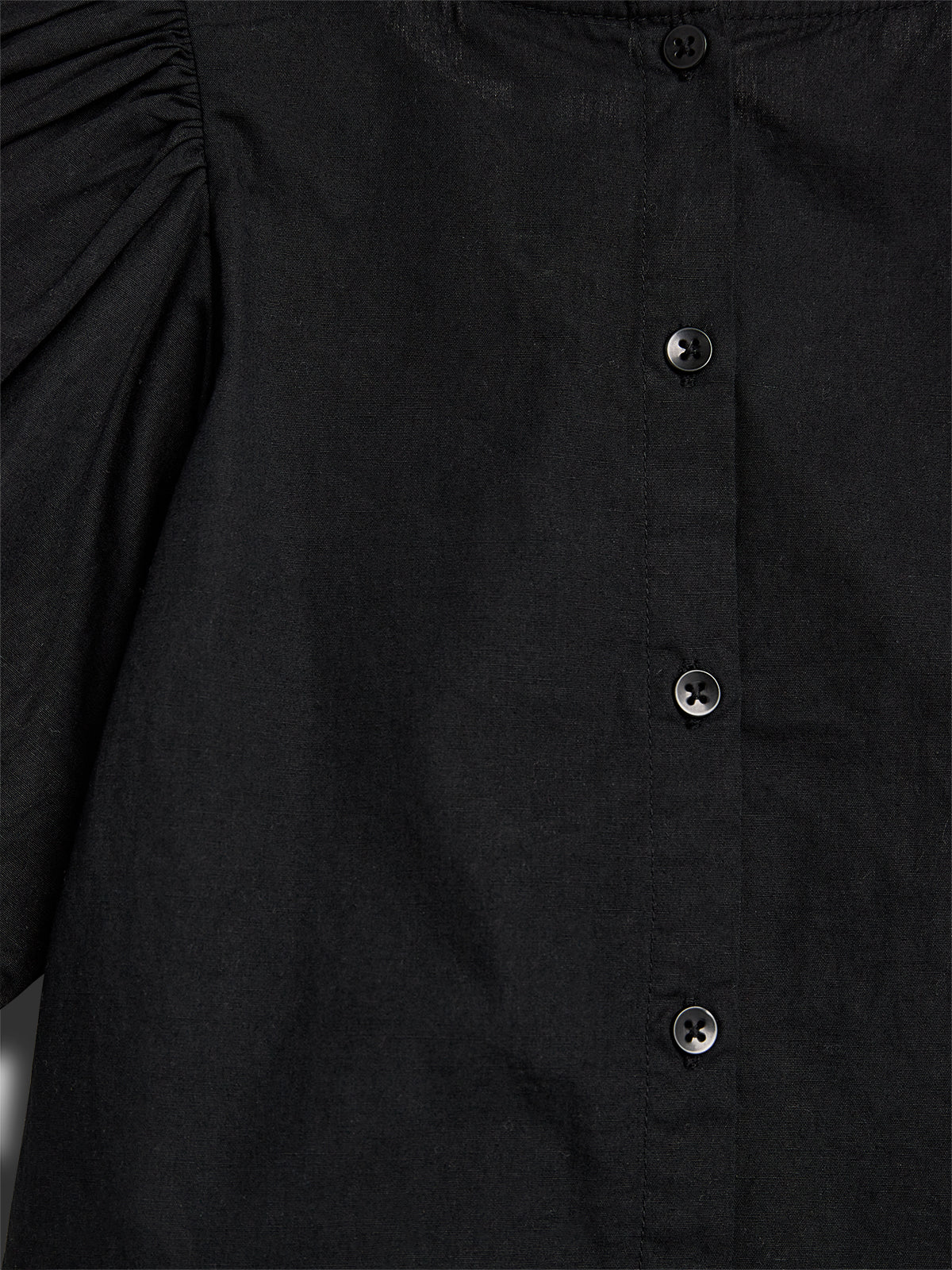 A black shirt features buttons down the front with ruched detailing on the shoulder, against a neutral background.