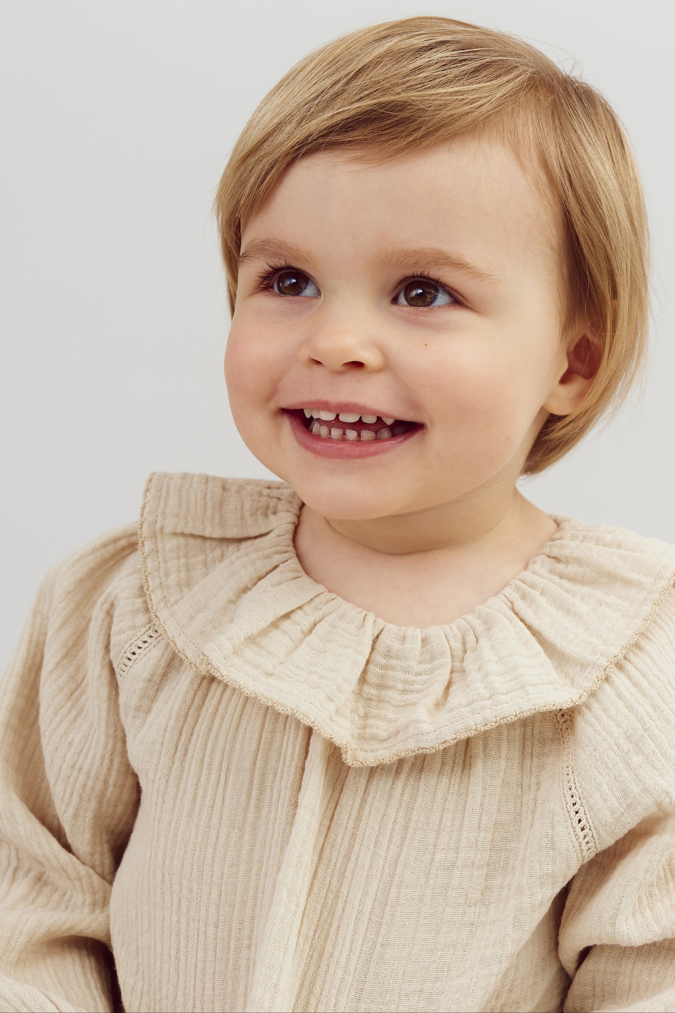 A toddler with brown eyes and short hair smiles, wearing a beige, ruffled, long-sleeve top. The background is plain and neutral, highlighting the child's joyful expression.