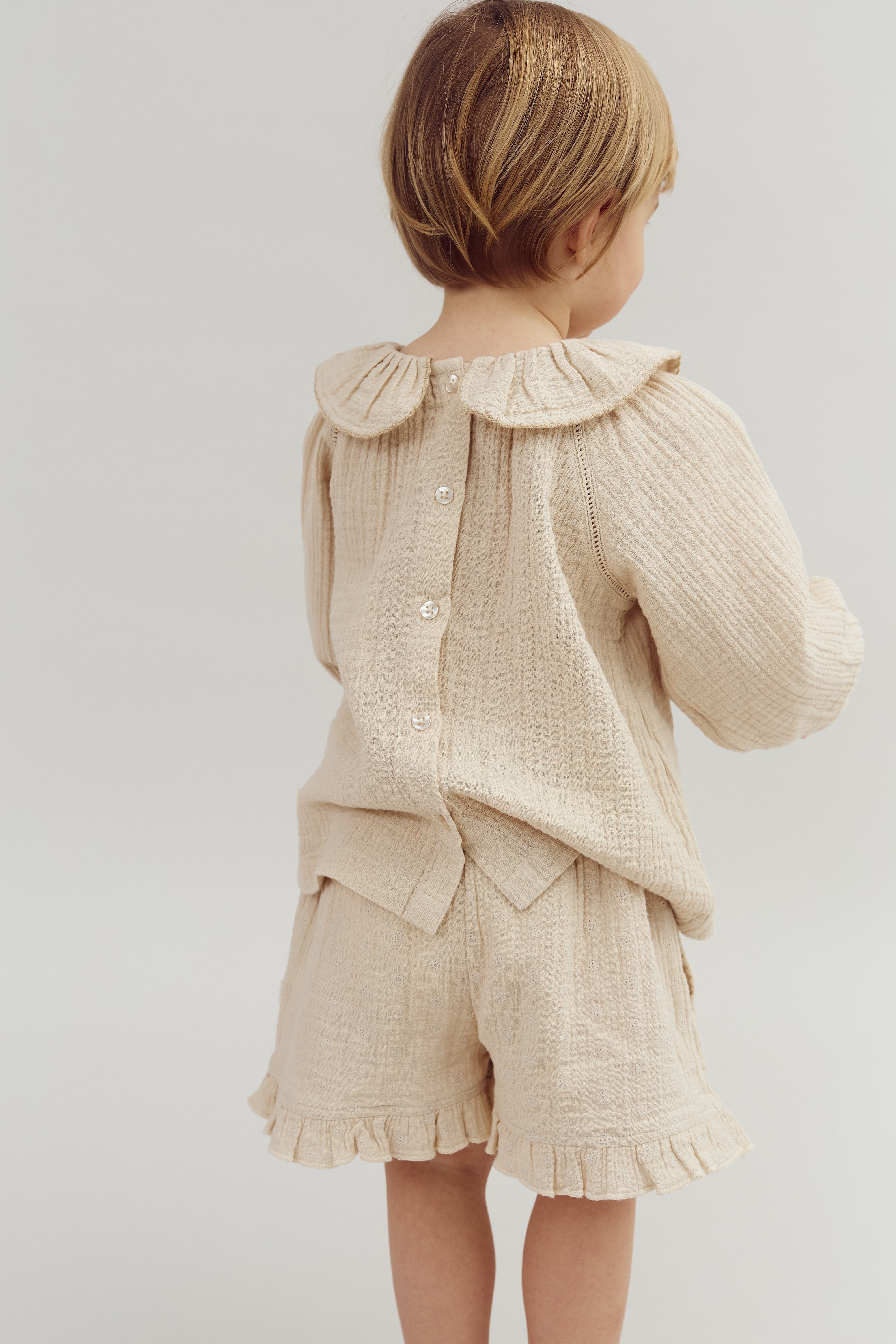 A child faces away, wearing a beige outfit with buttoned back and frilled edges, against a plain, light-colored background.