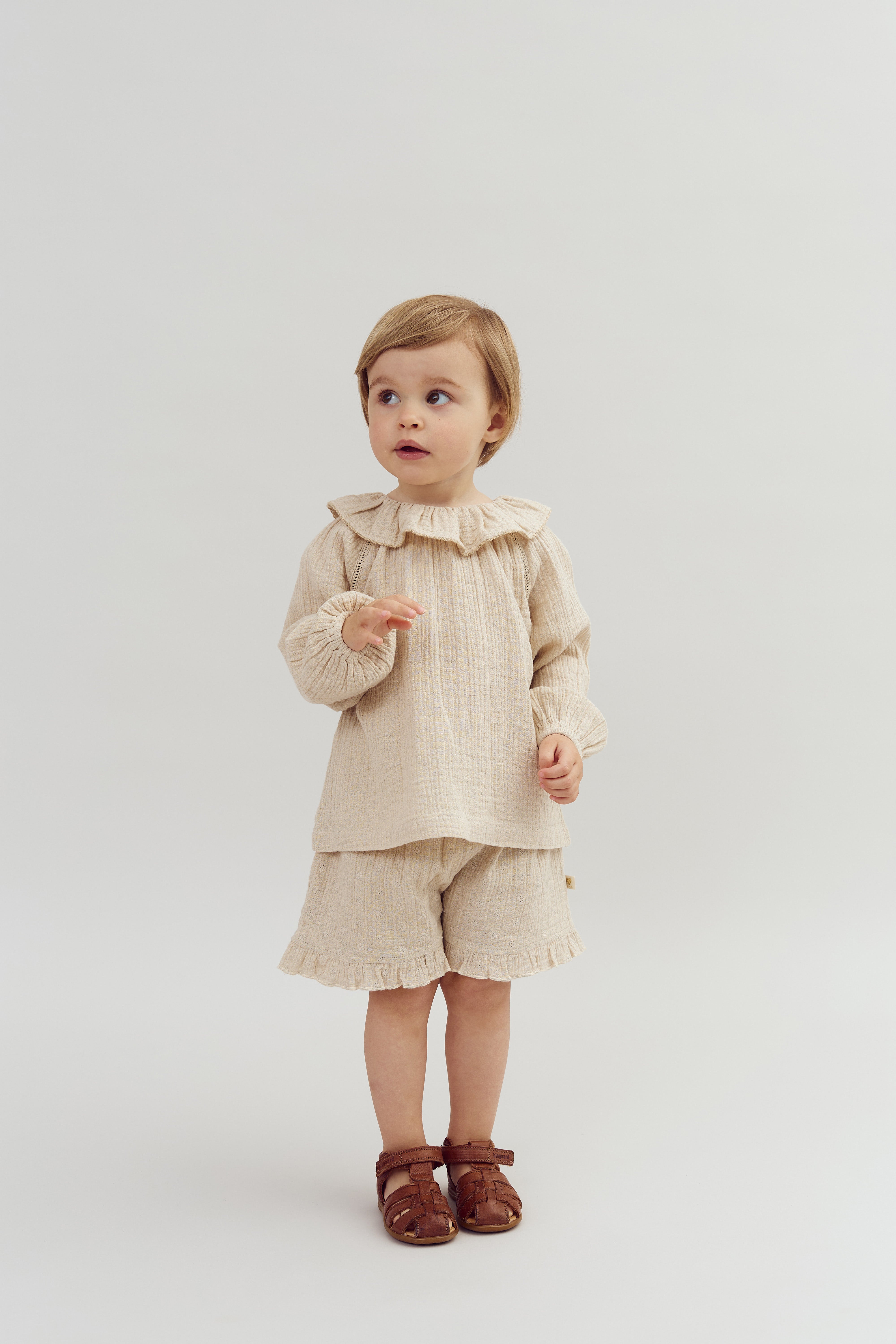 A young child wearing a beige outfit with ruffled collar and cuffs stands holding their hands mid-motion, in a plain, light-colored setting. They have short hair and are barefoot.