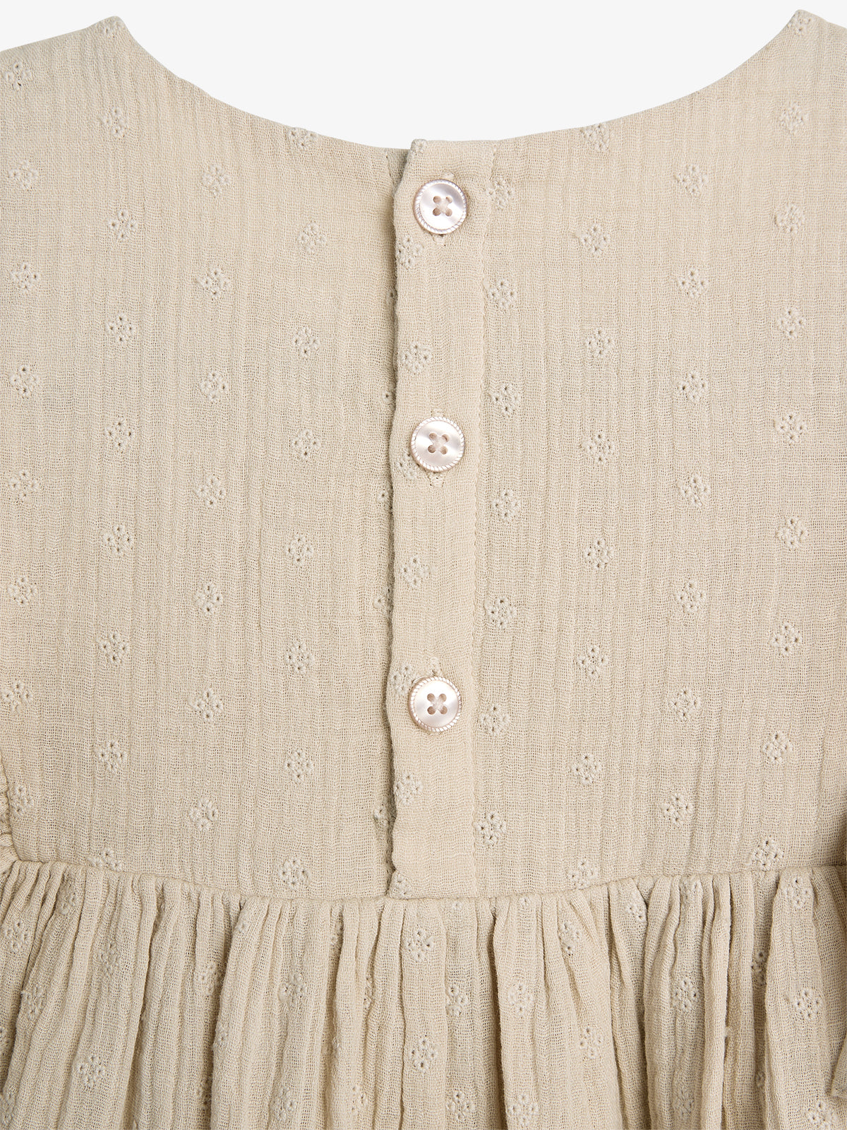 Beige dress back with three white buttons vertically aligned; delicate floral embroidery decorates the fabric. The dress features a gathered waist, highlighting a simple, elegant design against a plain background.