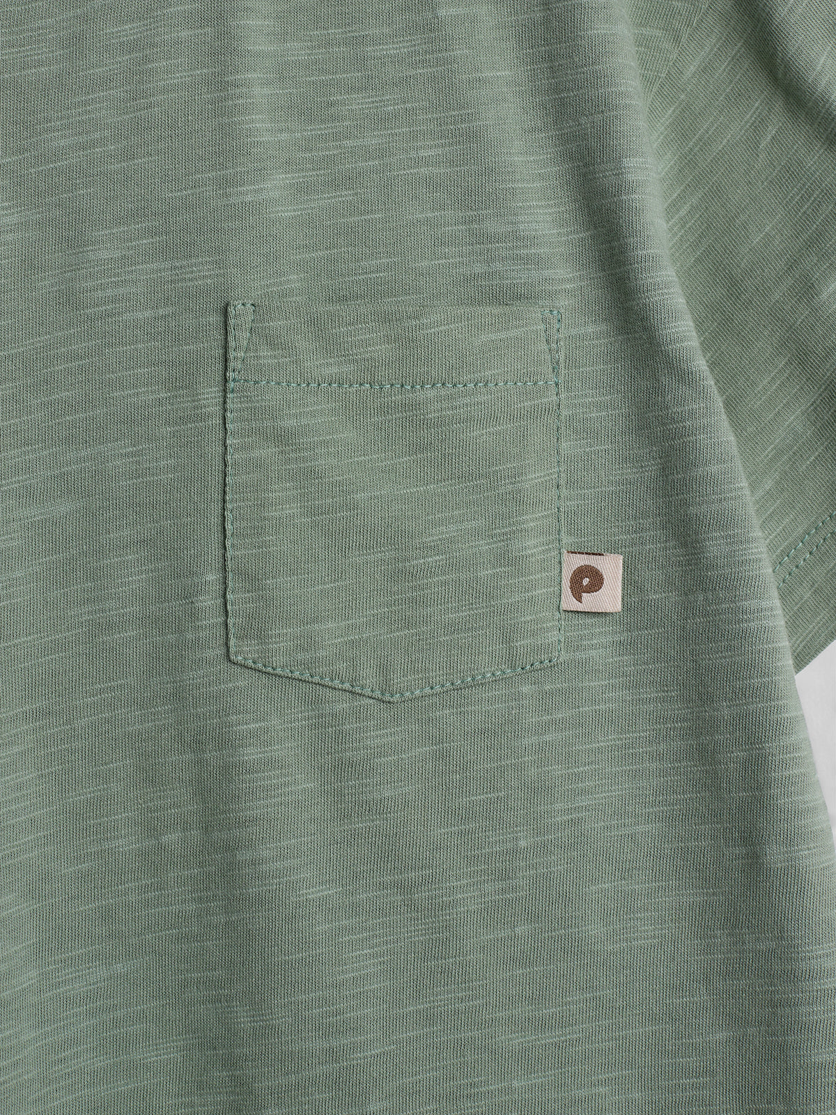 A green fabric with a small pocket stitched on it features a tiny tag with a simple dark brown design, set against a softly textured background.