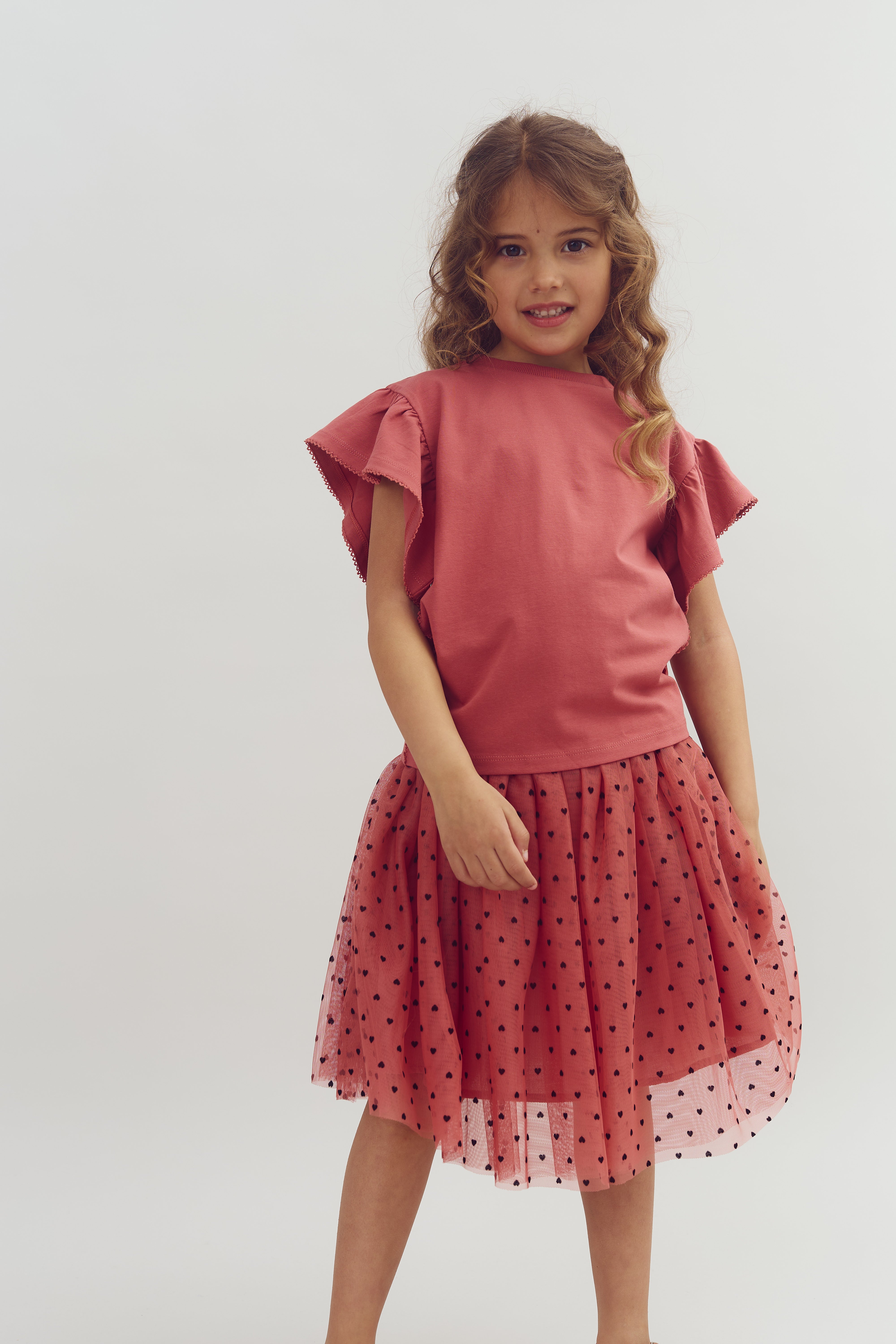 A young girl stands wearing a pink dress with flutter sleeves and a polka-dotted tulle skirt, smiling against a plain, light-colored background.