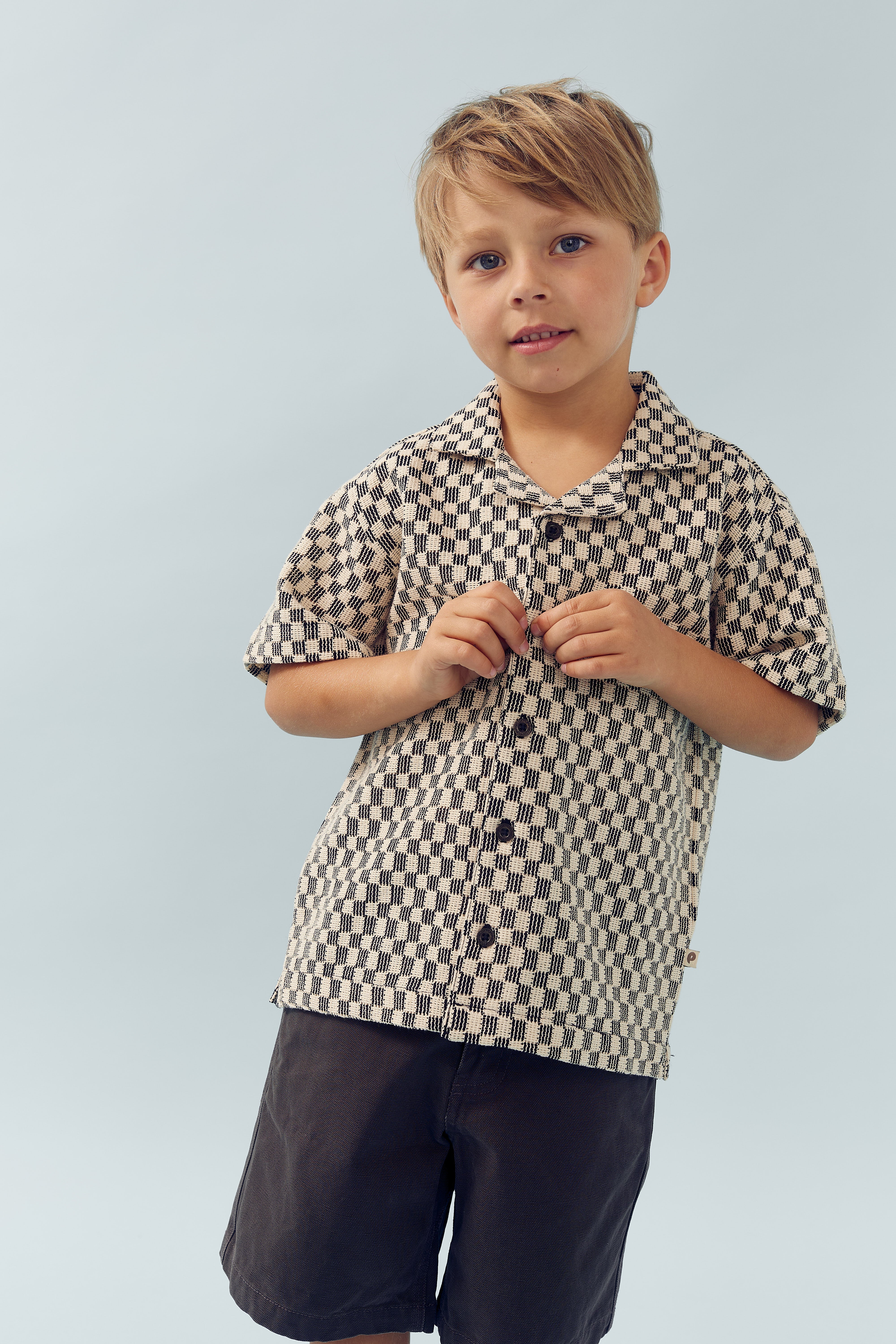 A young child, wearing a patterned button-up shirt and dark shorts, stands smiling slightly against a plain, light blue background.