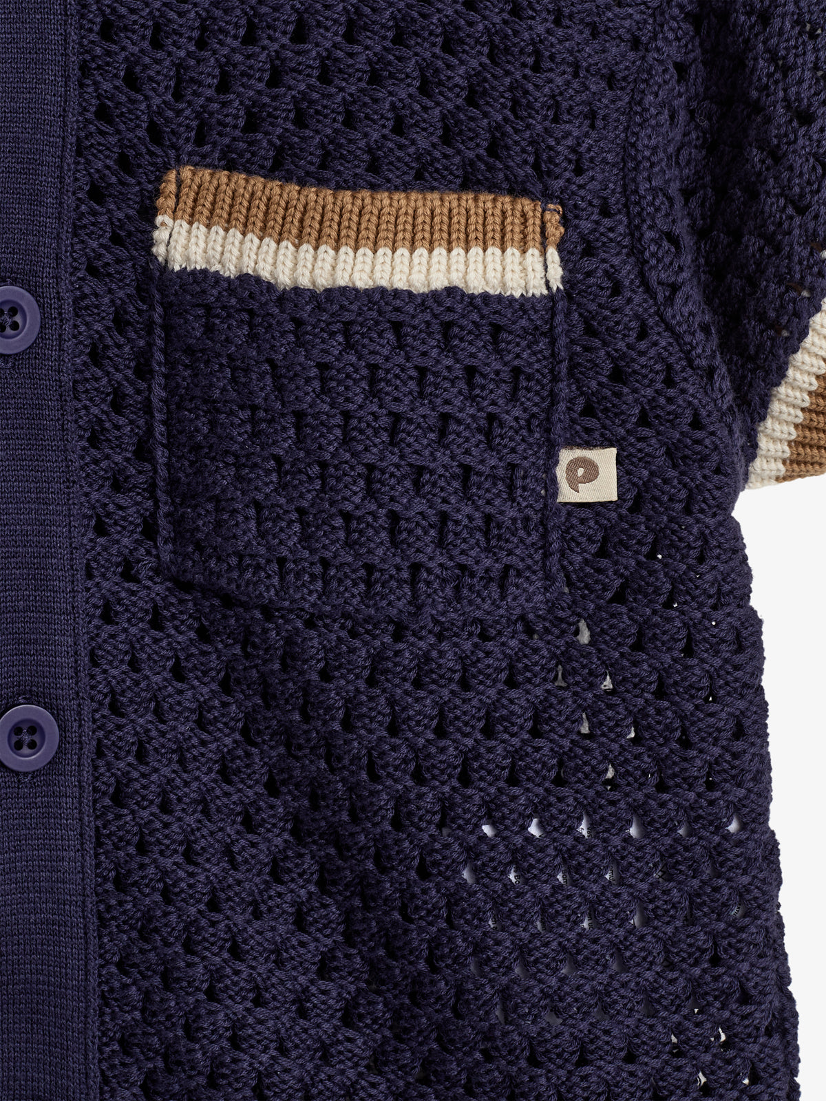 Cardigan med hæklet mønster i mørkeblå, har lilla knapper og en lomme med brune og hvide striber. Der er et lille mærke med bogstavet "P" ved lommen.