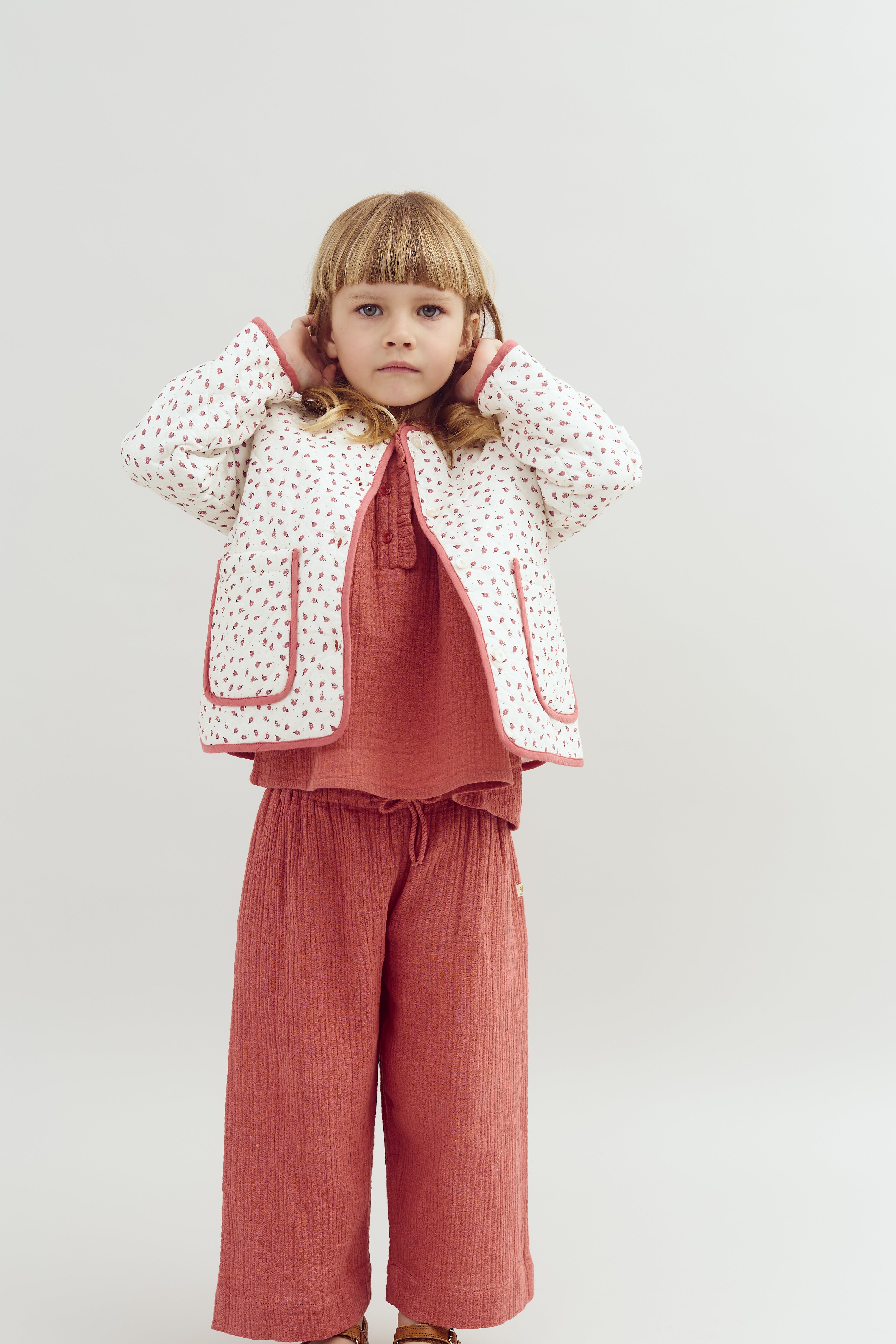 A young child stands with hands raised to their head, wearing a patterned jacket and pink outfit, in a neutral setting.
