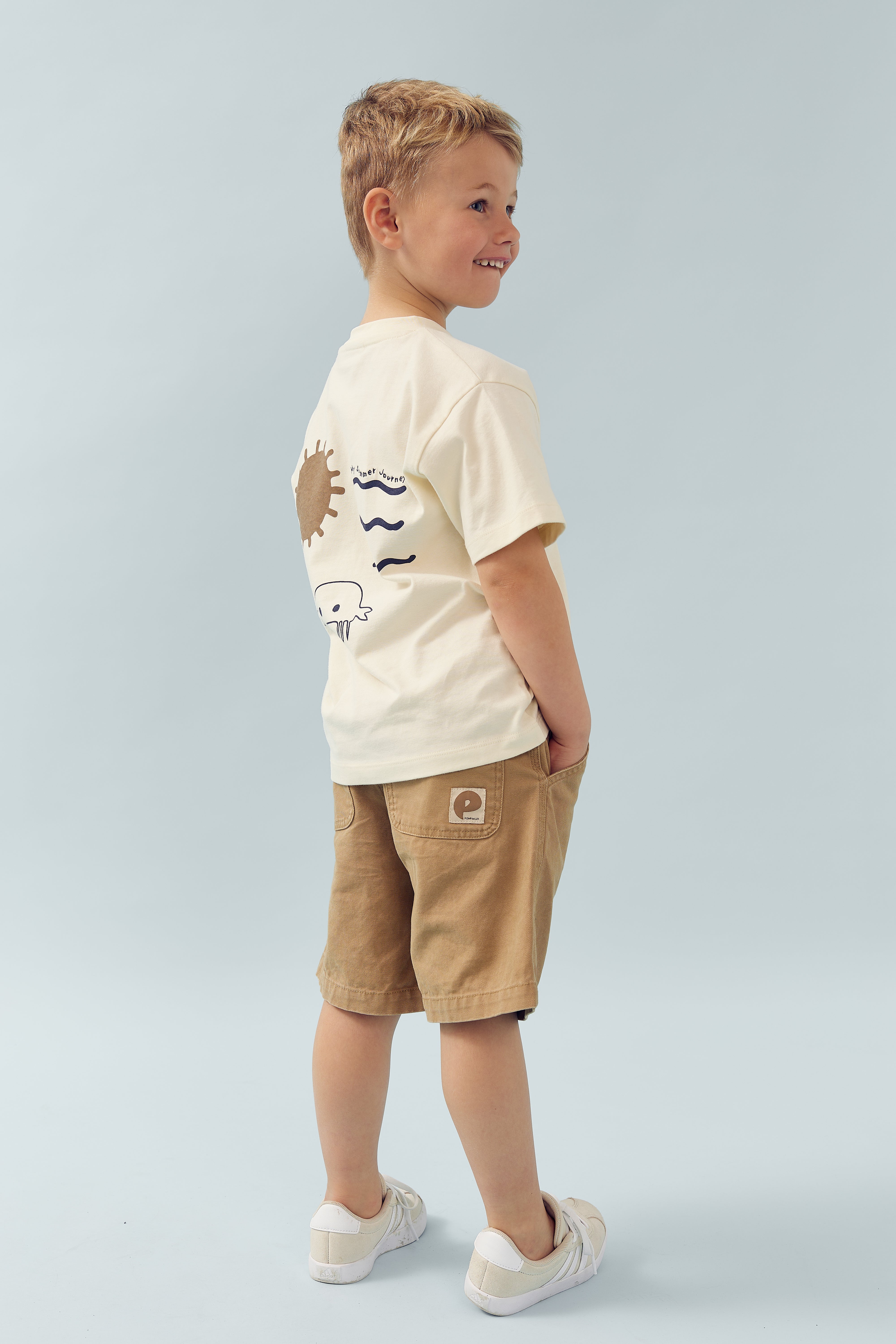 Boy in beige t-shirt and shorts stands smiling, with hands in pockets. T-shirt features sun and waves design. Bright, minimalist background.
