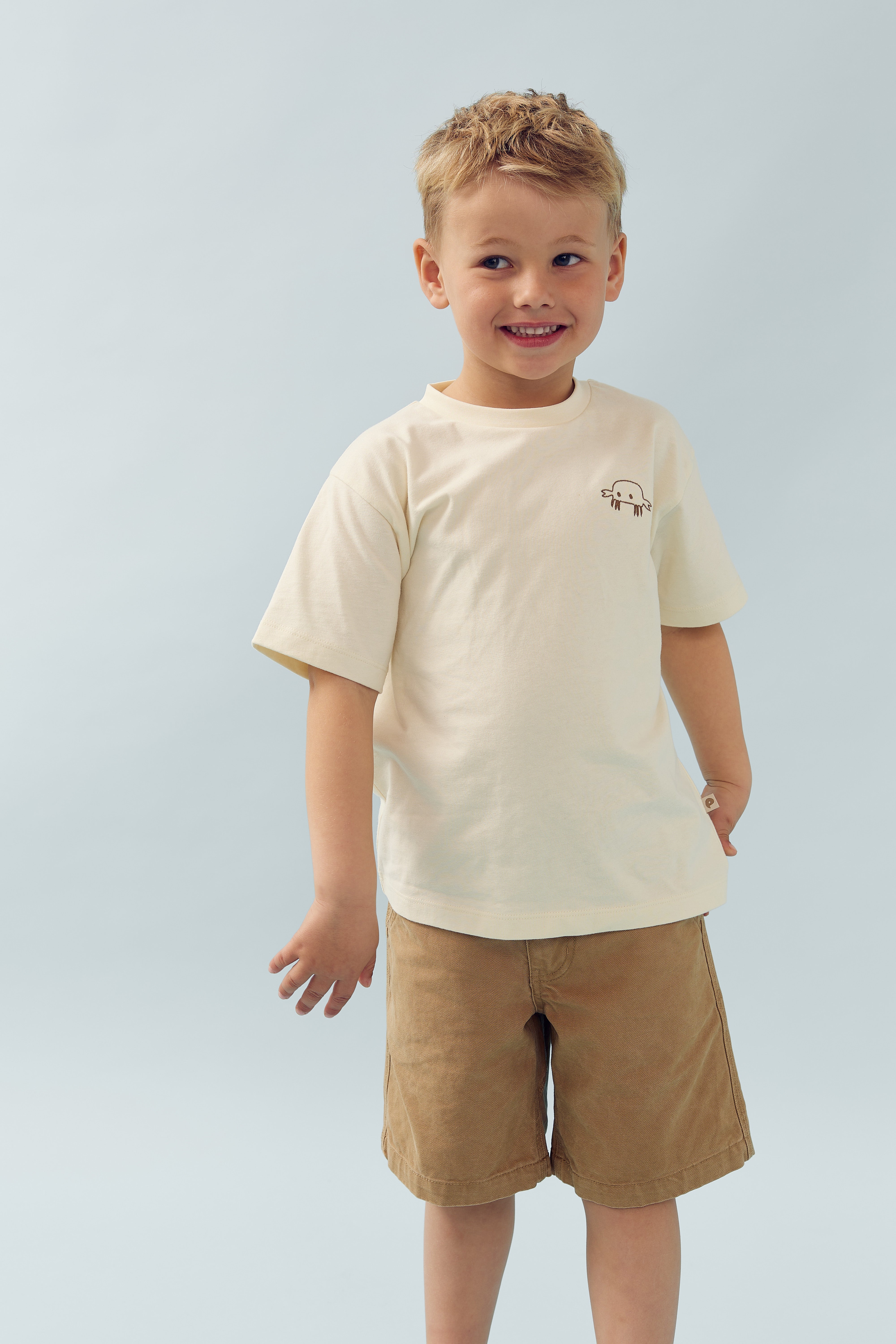 A young child smiles while standing, dressed in a cream T-shirt with a small jellyfish illustration and brown shorts, against a light blue background.