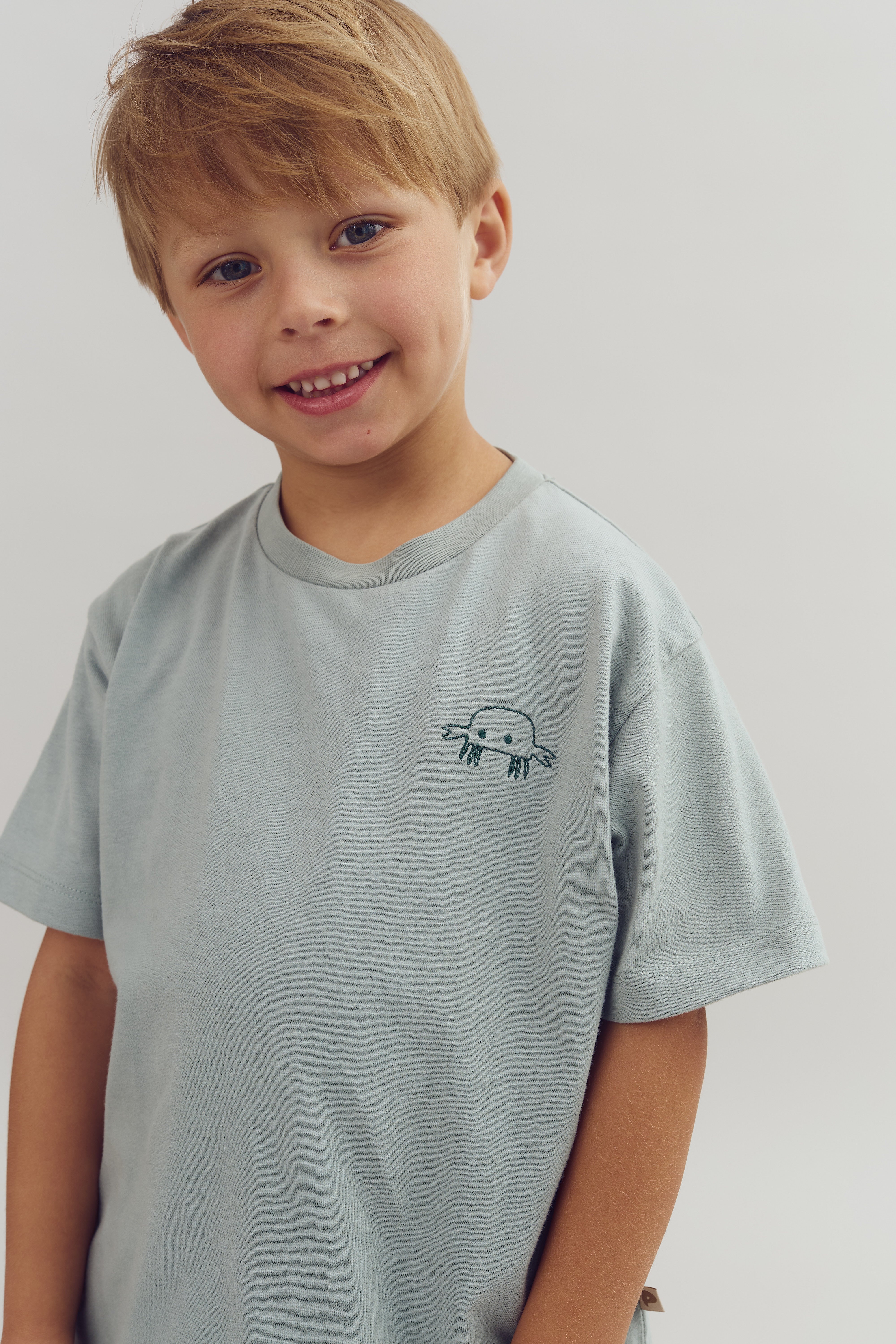 A child smiles, wearing a light gray T-shirt with a small cartoon jellyfish design on the chest, standing against a plain light background.