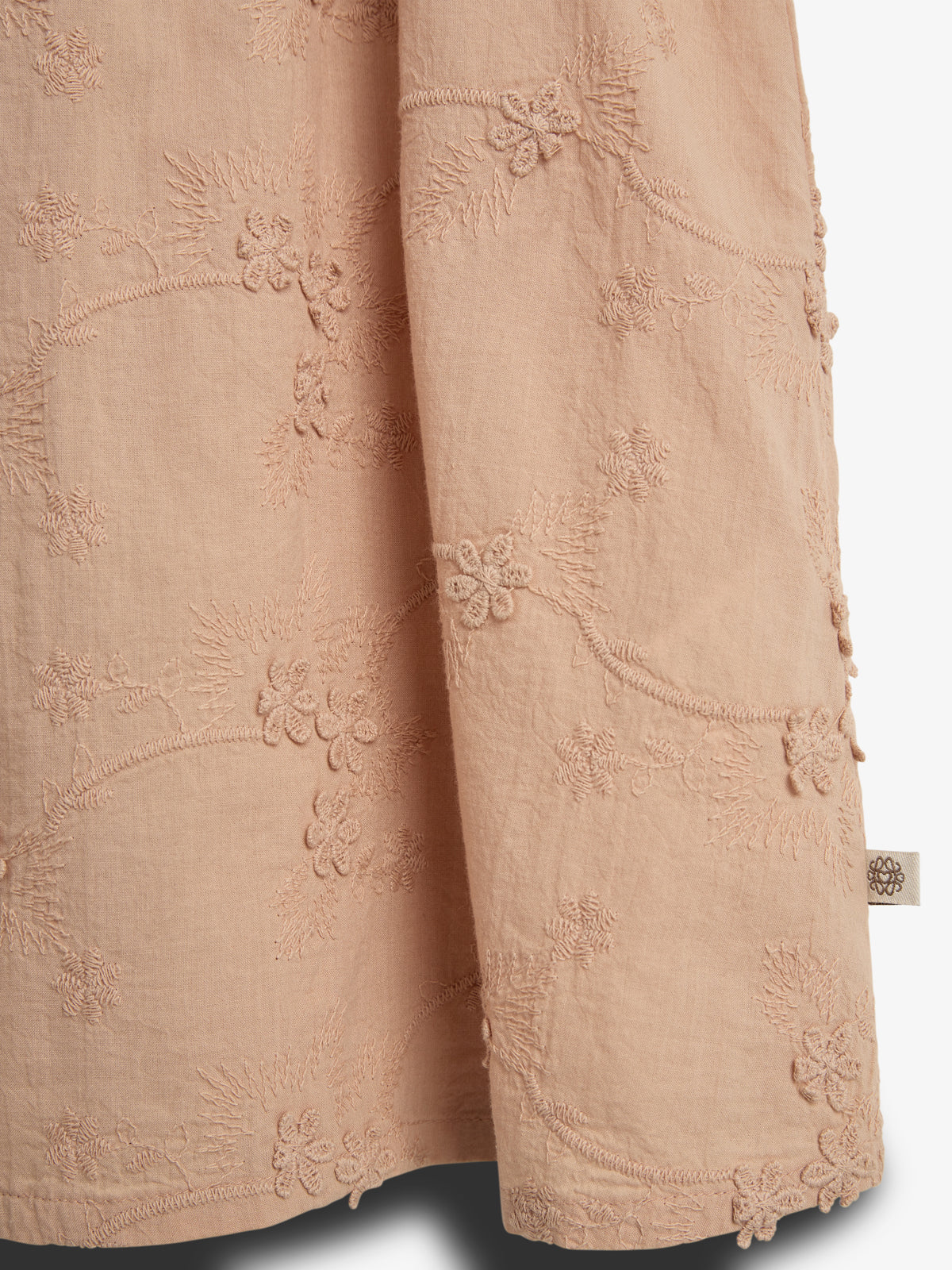 Beige fabric displays intricate floral embroidery forming patterns, with a small fabric tag featuring a floral logo. The background is plain, emphasizing the detailed designs on the textile.