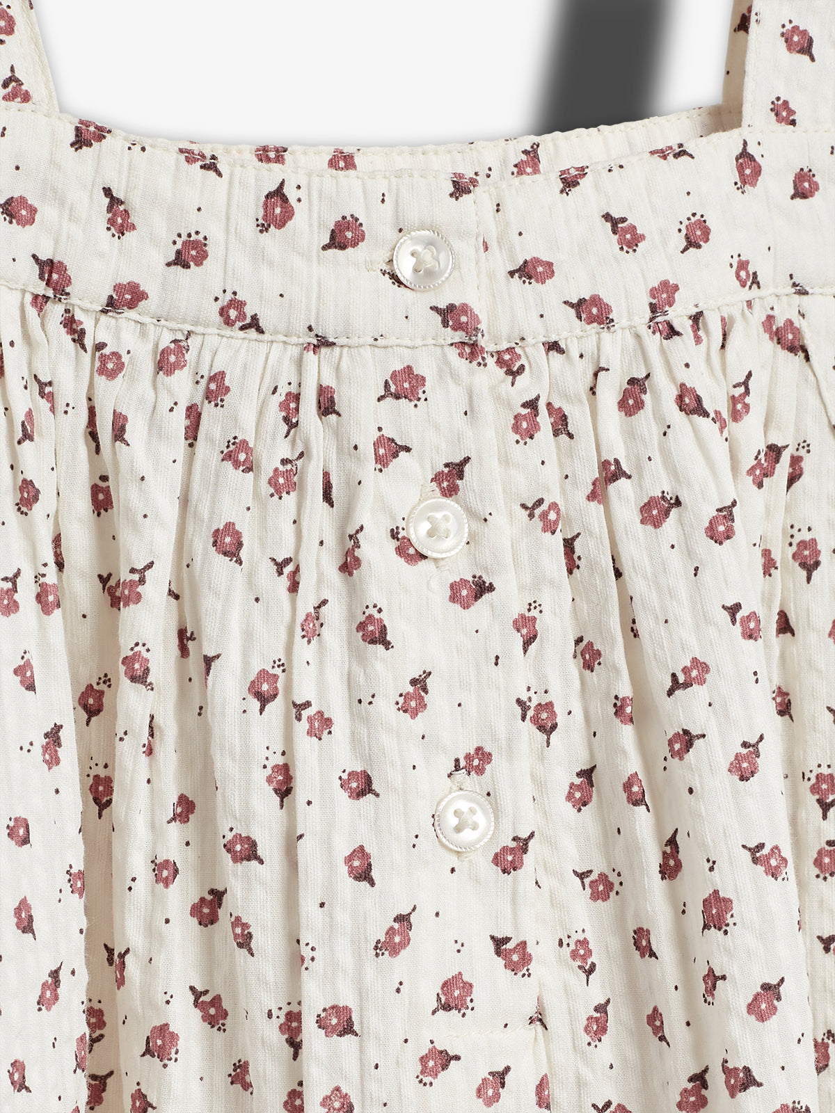 Kjole med små, røde blomstertryk og tre hvide knapper foran. Stoffet har en let krøllet tekstur. Baggrunden er lys og ensfarvet, hvilket fremhæver kjolens mønstre.