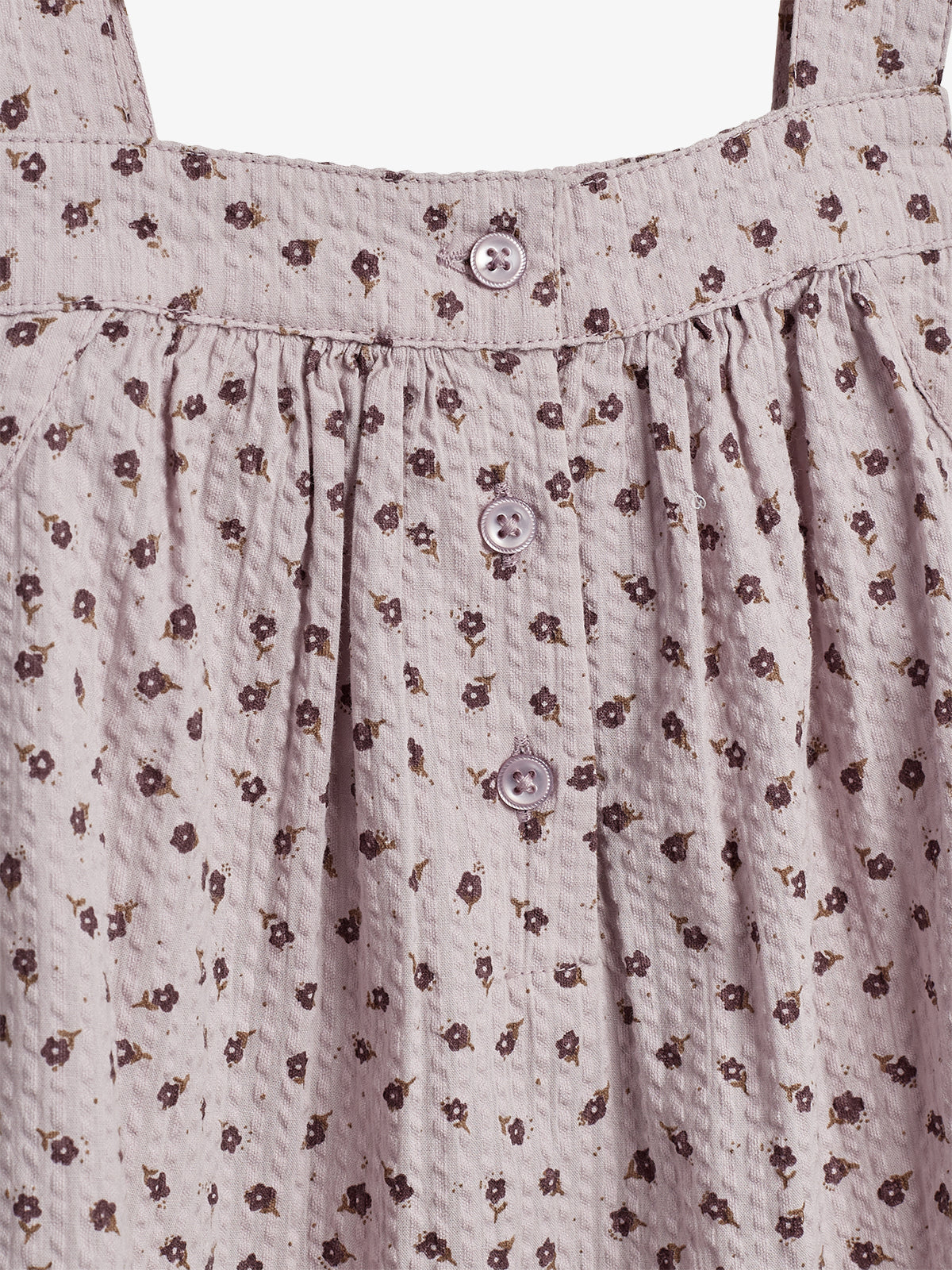 En lyslilla kjole med små, mørke blomsterprint og tre knapper på fronten. Stoffet har en krøllet tekstur, og der er stropper for oven.