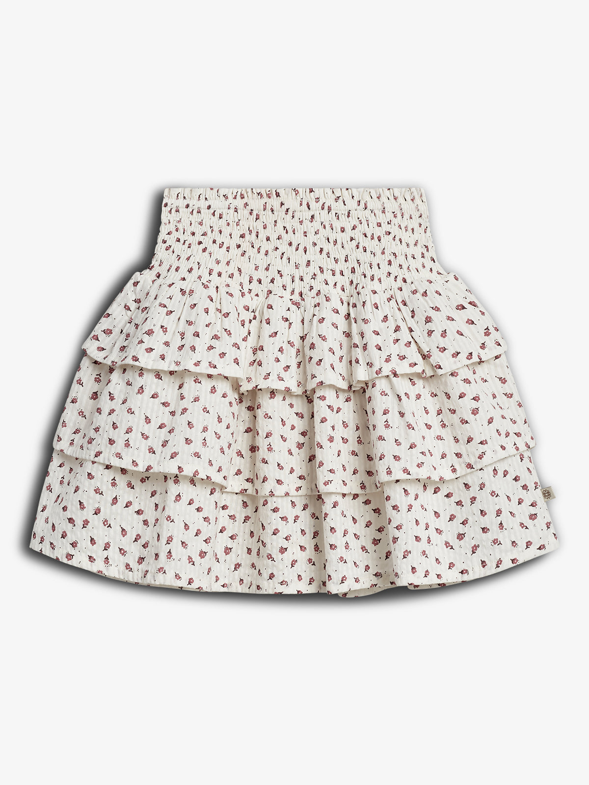 A white, tiered skirt with red floral patterns is displayed against a plain background. The skirt features a smocked waistband and three ruffled layers.
