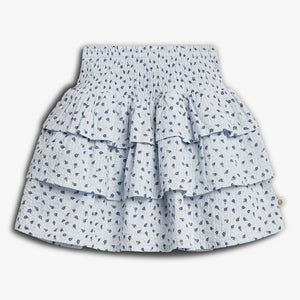 A light blue, tiered skirt with a floral pattern features small blue flowers. It has a smocked waistband and a layered, ruffled design, set against a plain white background.