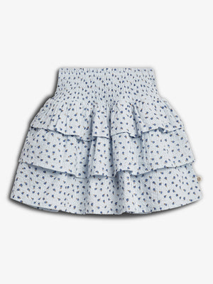 A light blue, tiered skirt with a floral pattern features small blue flowers. It has a smocked waistband and a layered, ruffled design, set against a plain white background.