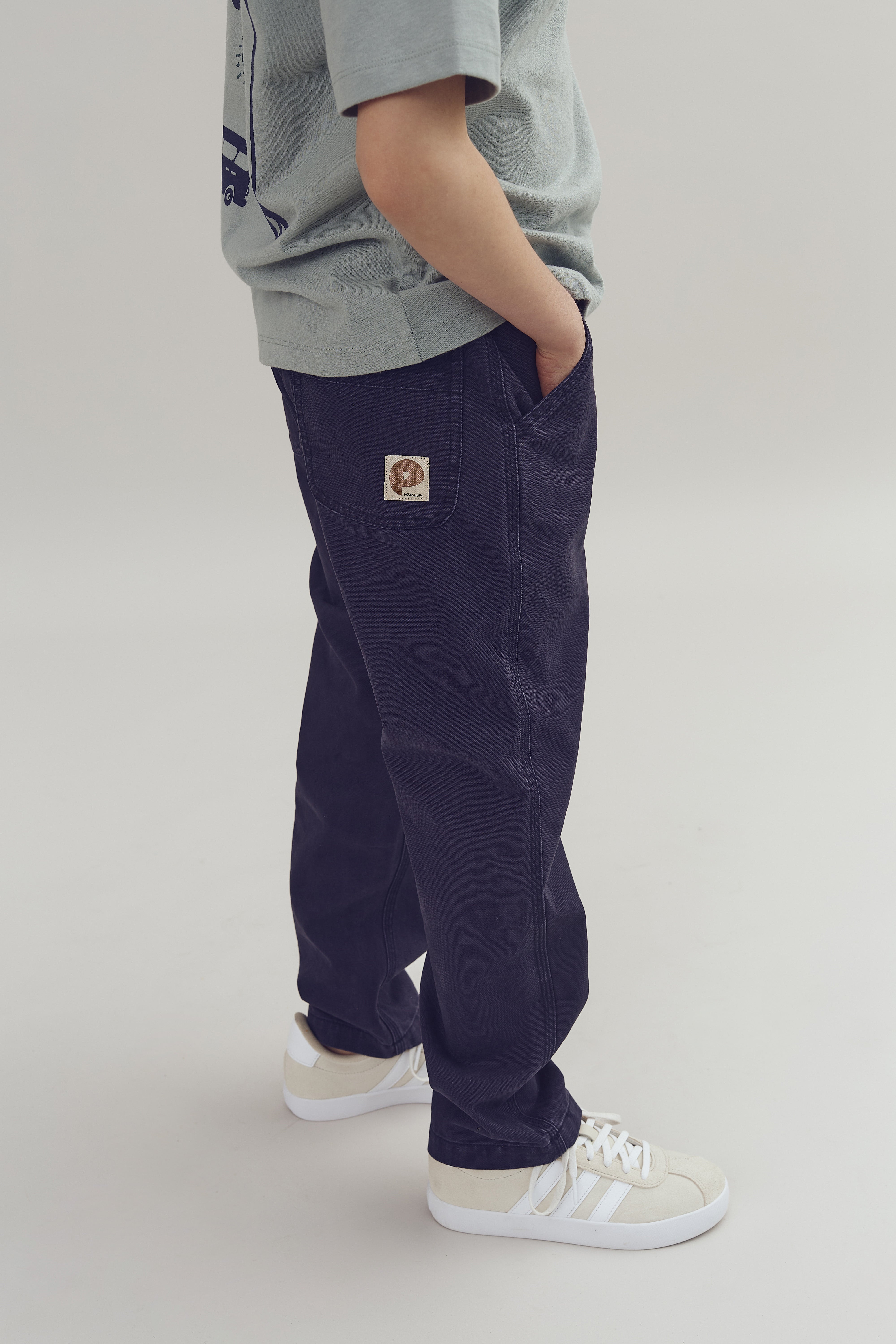 En person står iført en lysegrå T-shirt, mørke jeans med en lomme med "p" logo, og hvide sneakers. Baggrunden er ensfarvet lys grå.