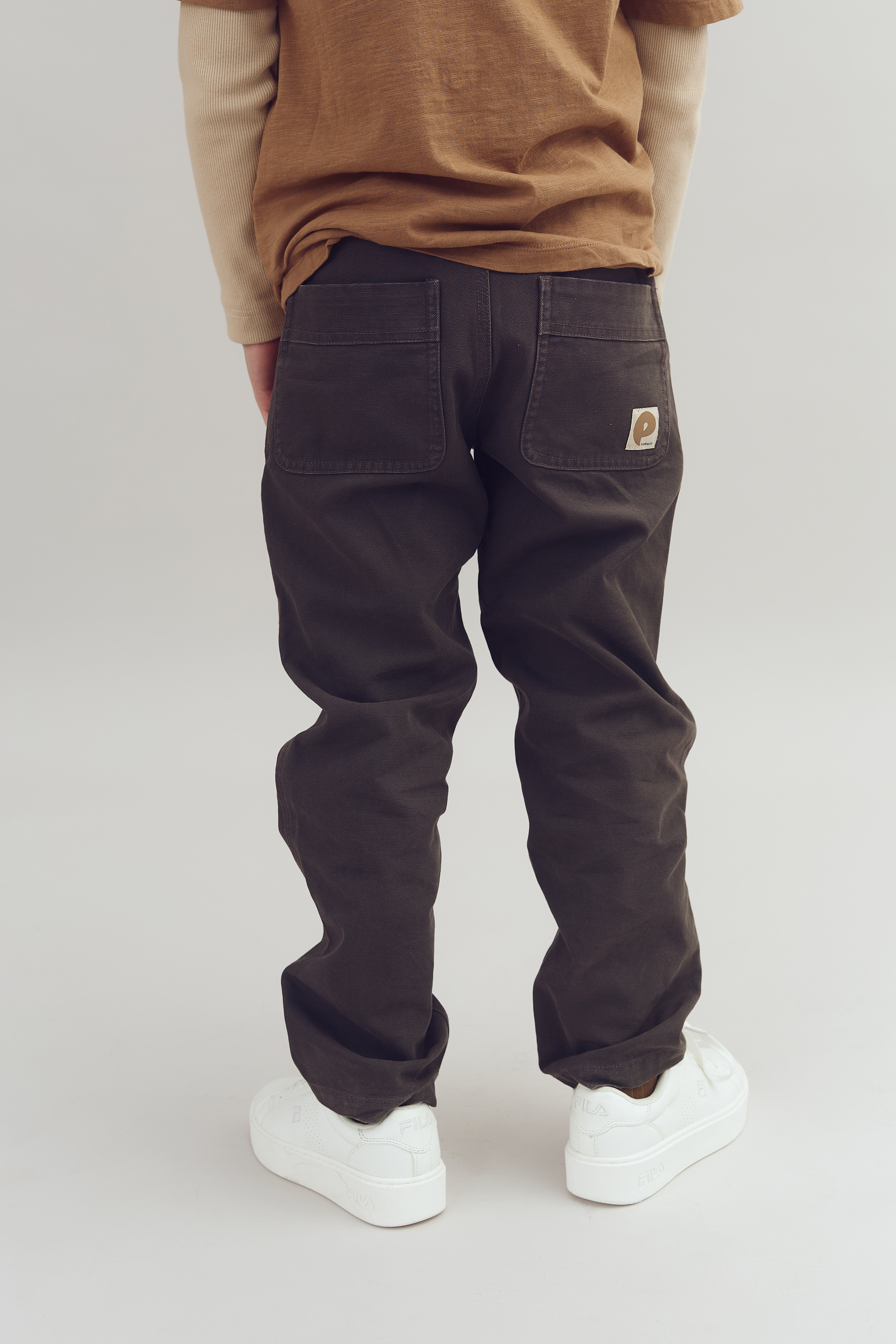 Person iført brune bukser med to baglommer og hvid tekst på venstre samt hvide sneakers. Bukserne bæres afslappet. Baggrunden er en neutral, lys farve.