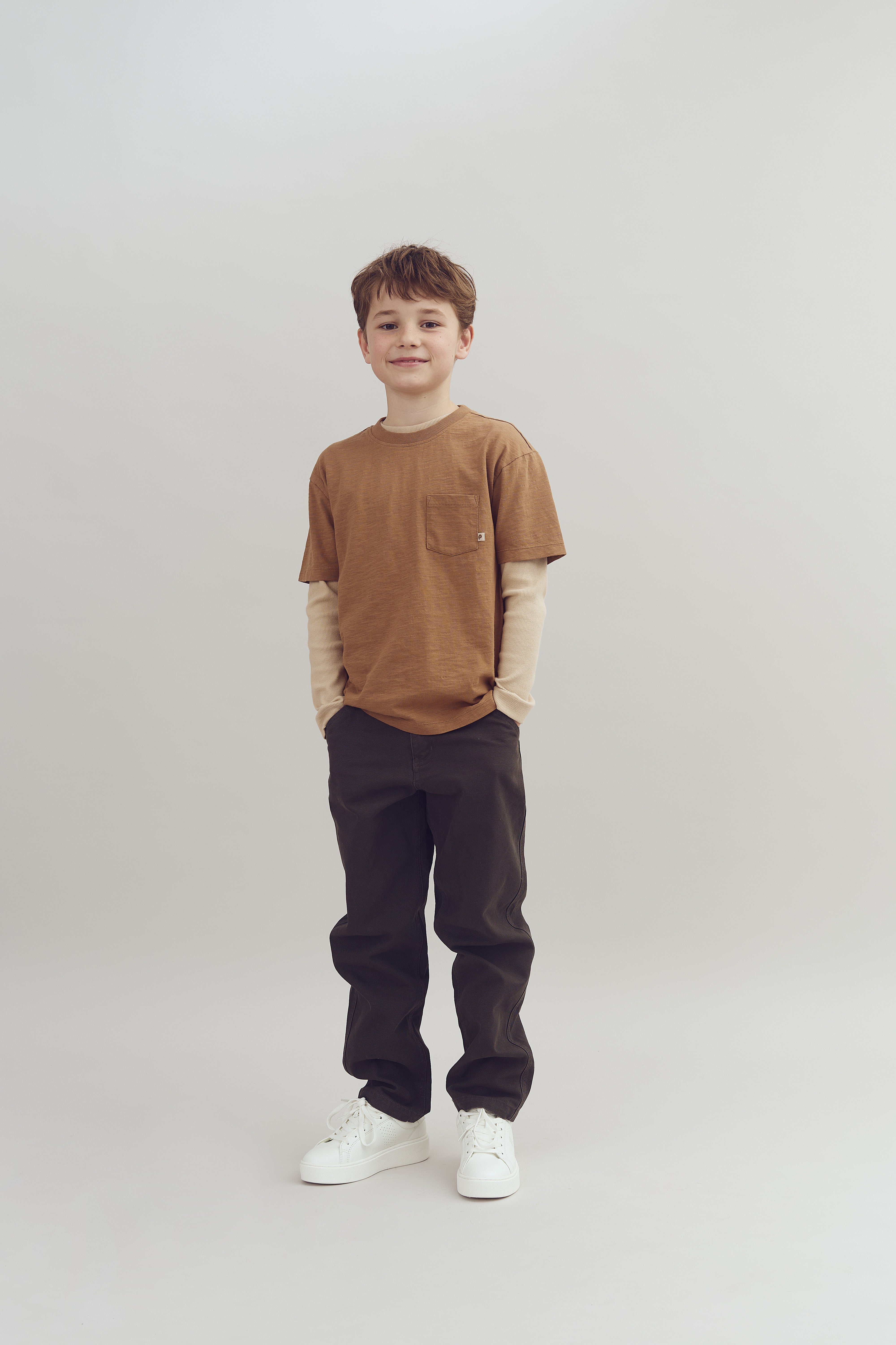 Dreng står stille, iført en brun t-shirt, lange ærmer, mørke bukser og hvide sneakers. Baggrunden er neutral og lysegrå.