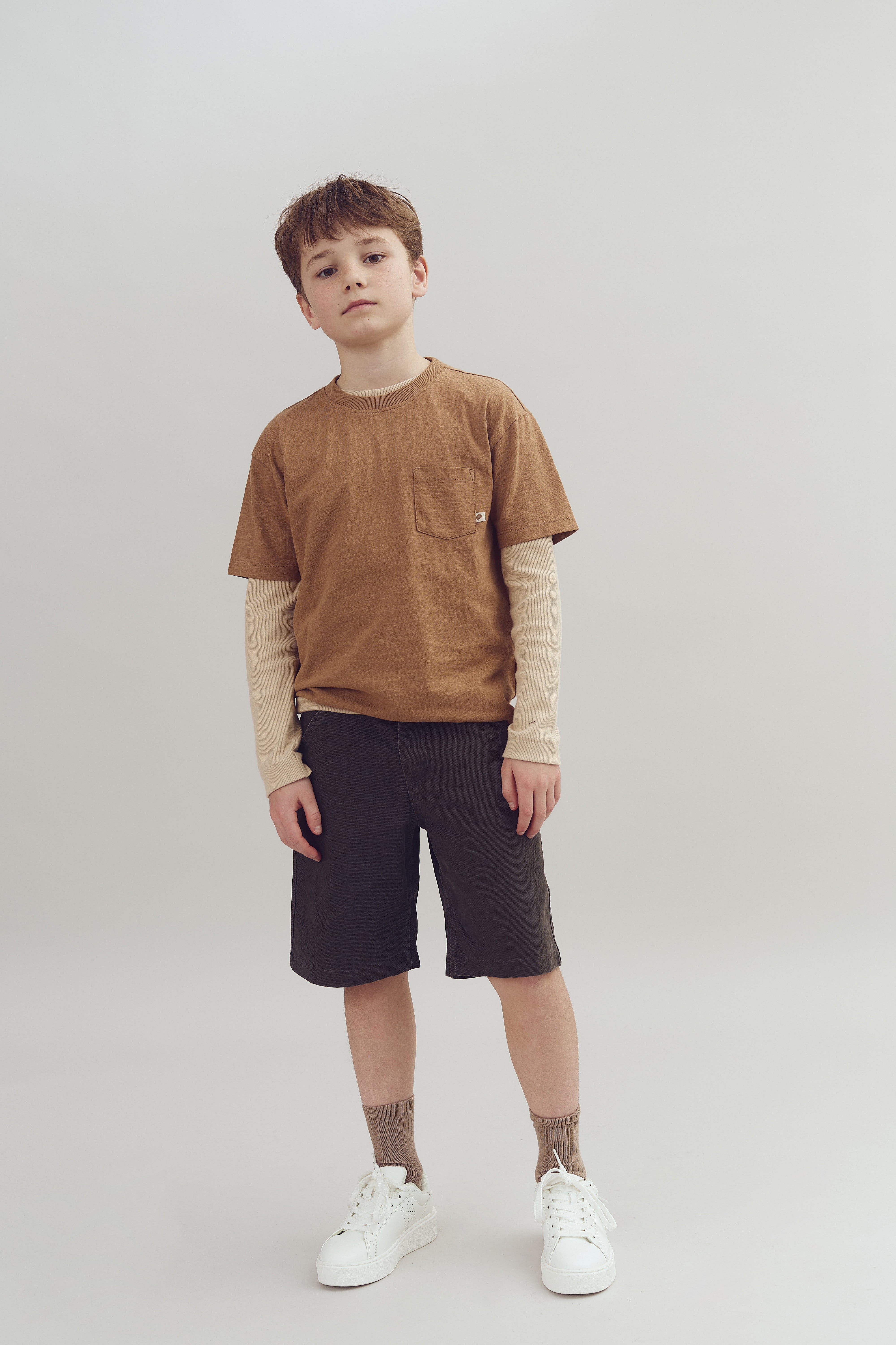 Dreng stående afslappet, iført brun T-shirt, lange ærmer og mørke shorts, på en simpel, lys baggrund. Han bærer hvide sneakers og ser direkte fremad.