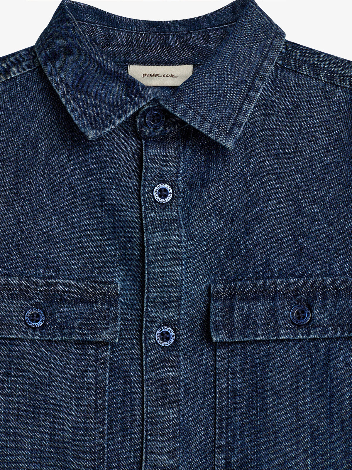 En denimskjorte med krave og knapper i forgrunden. Skjorten har to brystlommer med knapper. Etikettekst: "POMPdeLUX". Skjorten er tilsyneladende på en neutral, lys baggrund.