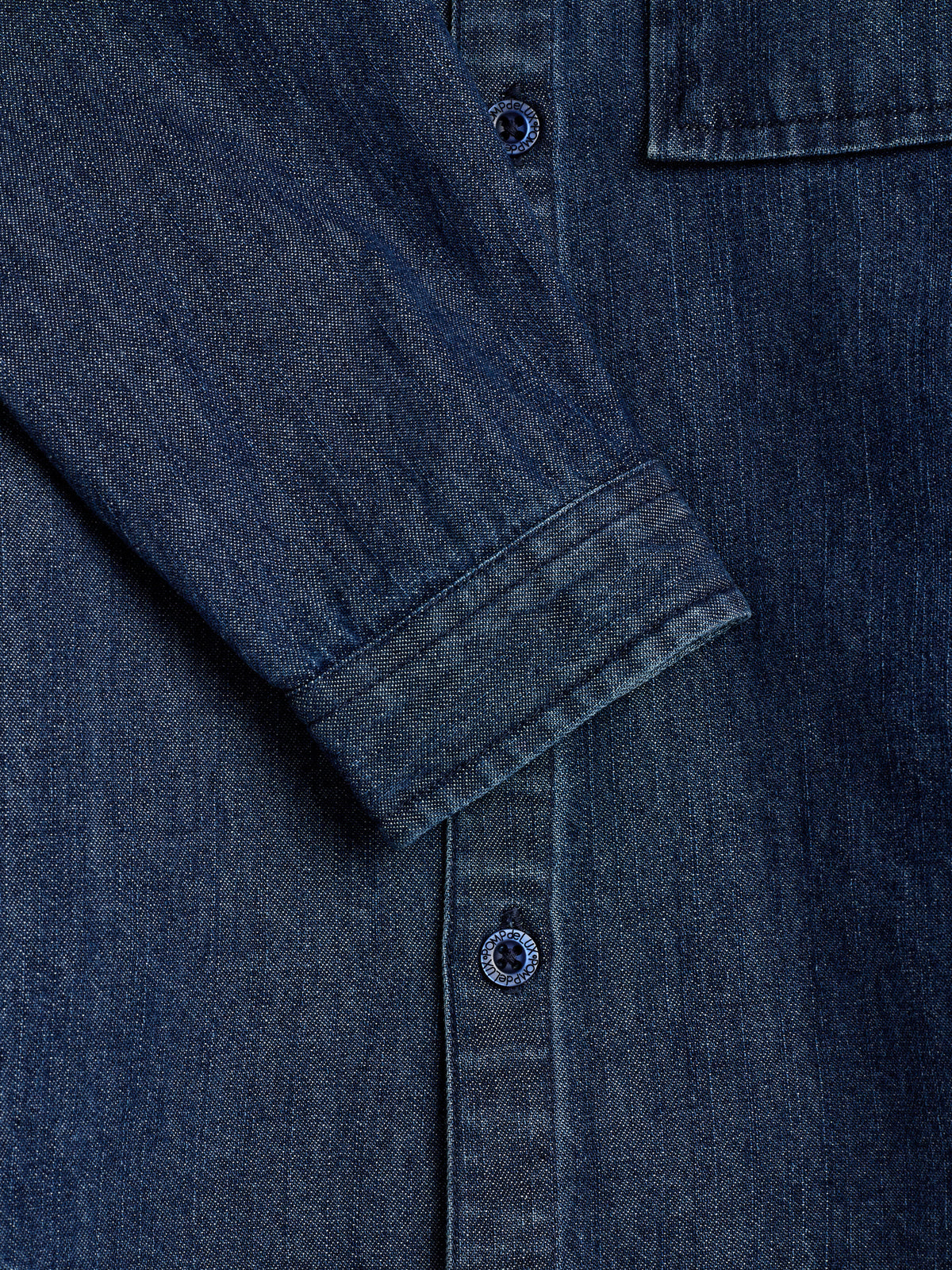 En denimskjorte ses tæt på med en knappet manchet og to knapper synlige langs lukningen. Stoffet er blåt og teksturen er tydelig.