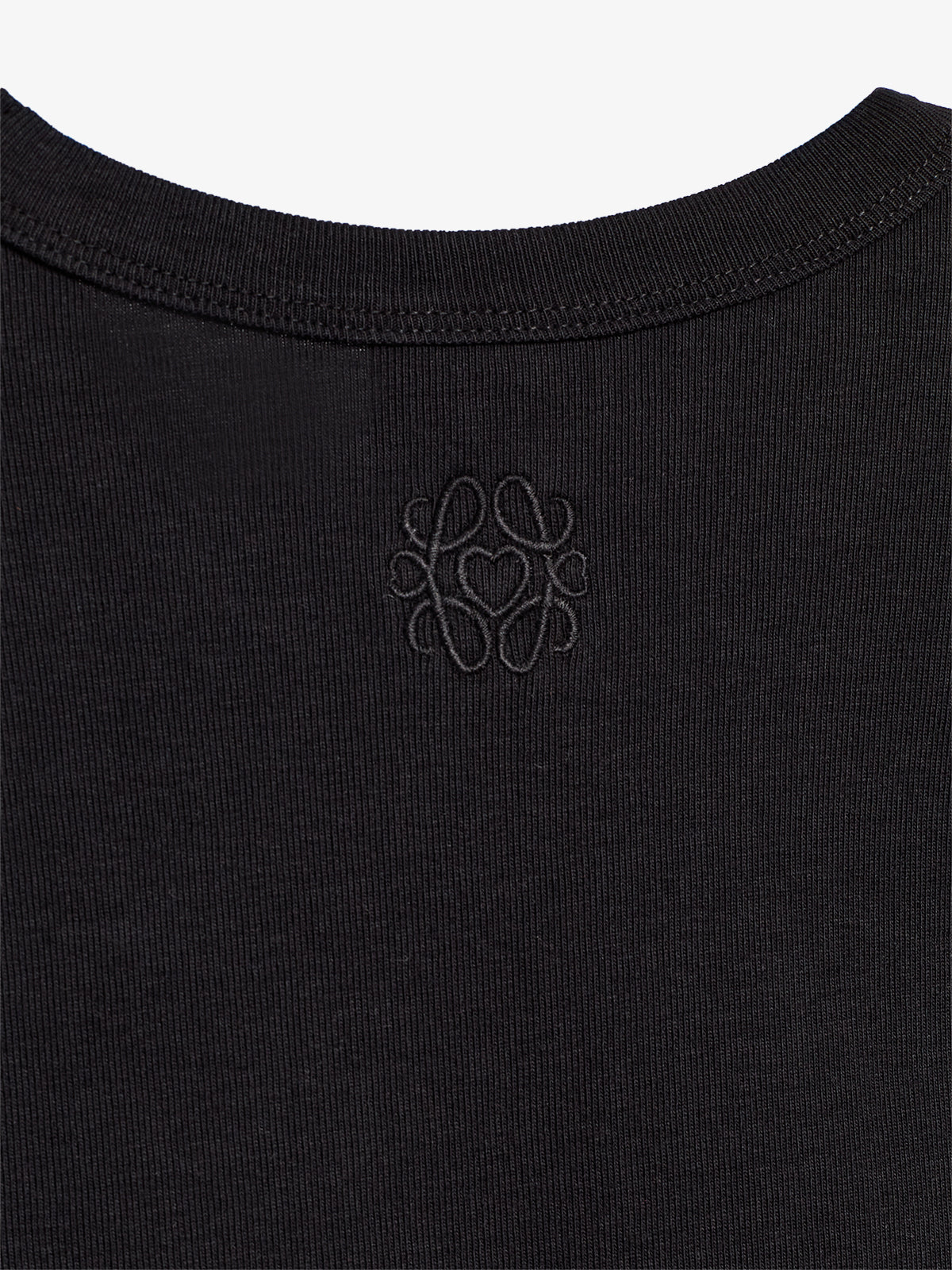 En sort T-shirt viser en diskret, broderet logo ved nakken. Baggrunden er ensfarvet, hvilket fremhæver det detaljerede mønster.