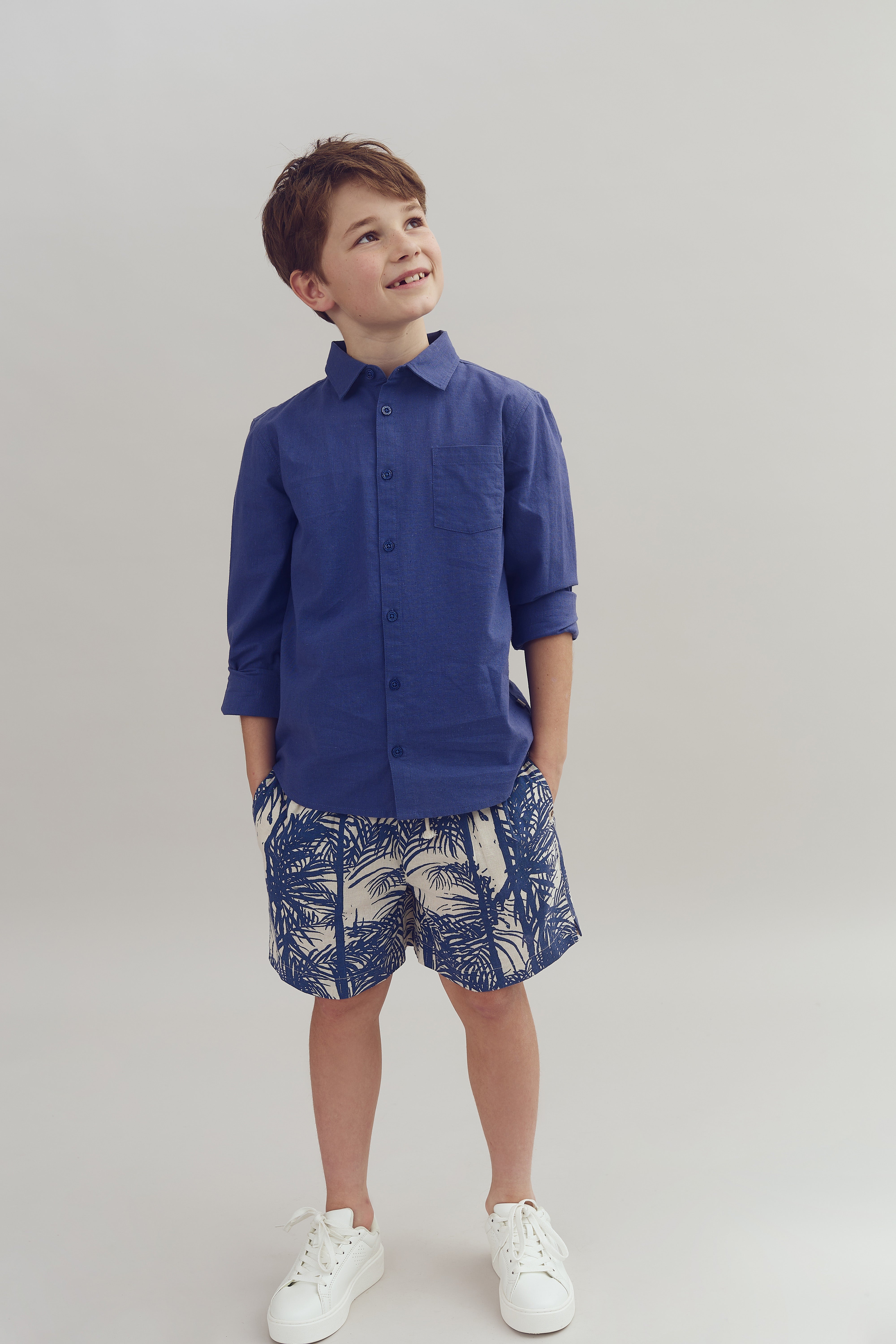 Dreng smiler, iført blå skjorte, mønstrede shorts og hvide sneakers, i neutralt oplyst rum.