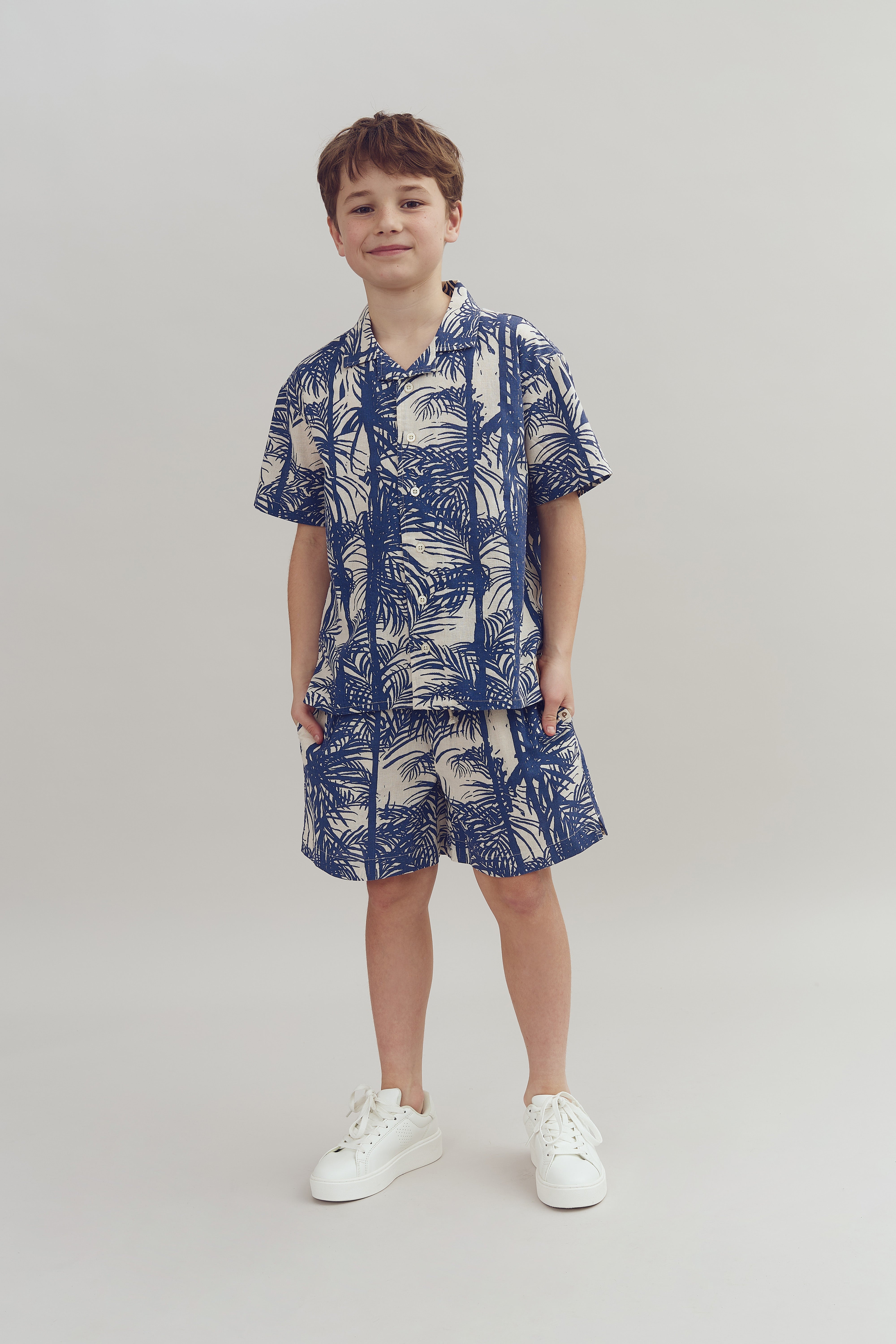 Dreng iført en blå-hvid mønstret skjorte og shorts står stille, smilende, med hænderne i lommerne. Han har hvide sneakers på, og baggrunden er neutral.