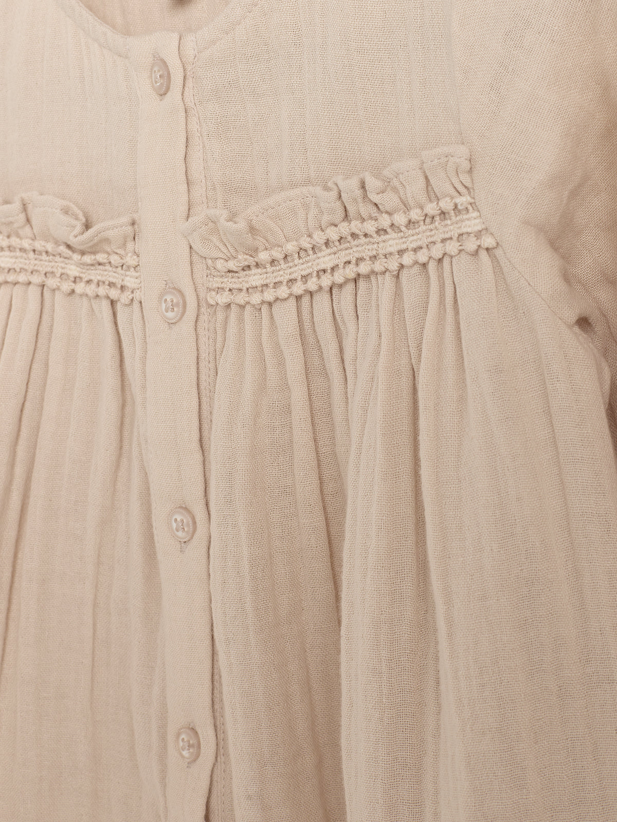 Beige bluse viser detalje med knapper og rynkede, dekorative folder øverst. Stoffet er let og struktureret, velegnet til afslappet brug. Ingen tekst er synlig.