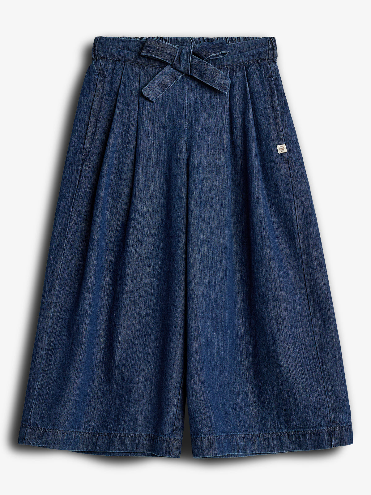 Et par vide, mørkeblå denim culotte-bukser ligger fladt. Bukserne har et bindebånd i taljen og stiklommer, med en lille firkantet mærkat på højre side. Baggrunden er hvid.