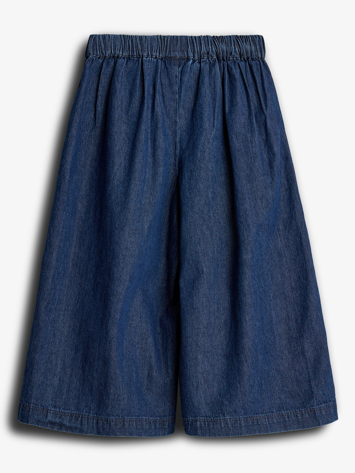Blå denimnederdel med elastisk talje, hænger i folder og falder løst omkring. Baggrunden er ensfarvet hvid, hvilket fremhæver nederdelens struktur og materiale.